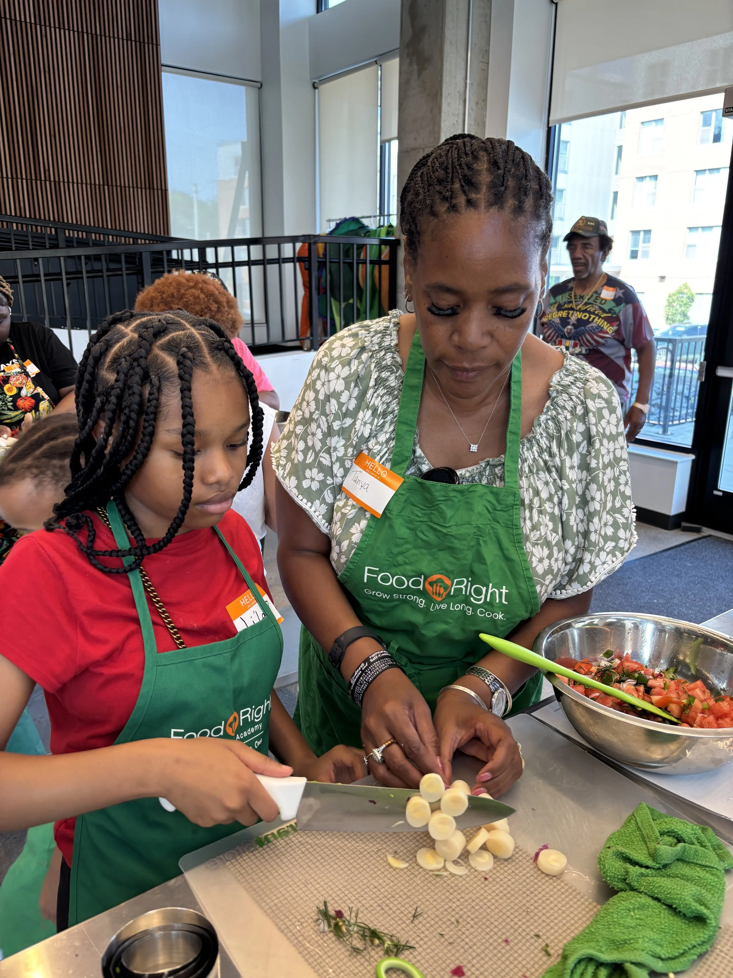 A woman and a girl chop vegetables in a kitchen, with a large bowl of salad on the counter, wearing green aprons with 'FoodRight' logo.