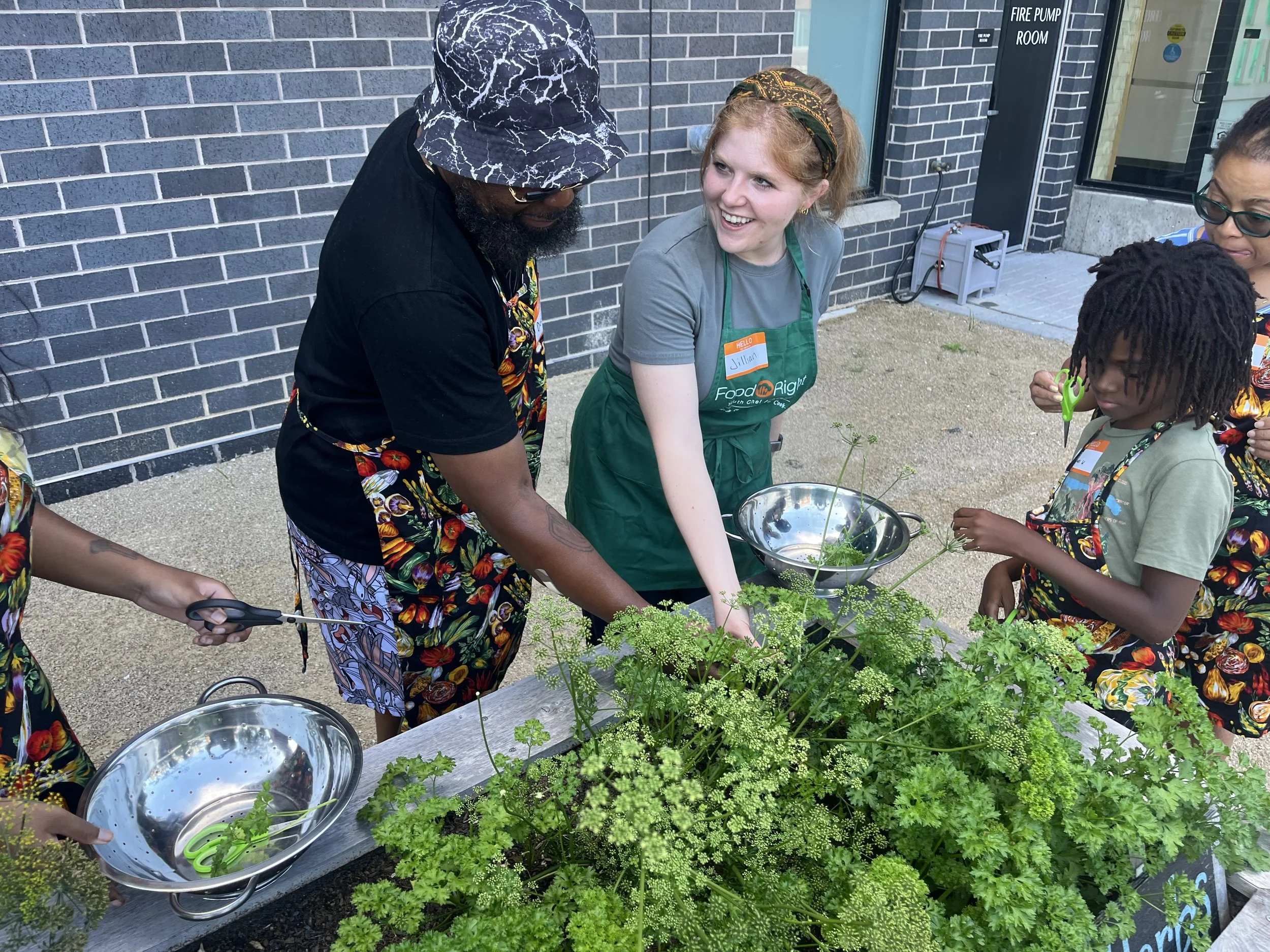 A group of people, including women and children, gathered outdoors near a brick building, participating in a gardening activity with green plants and herbs on a table.