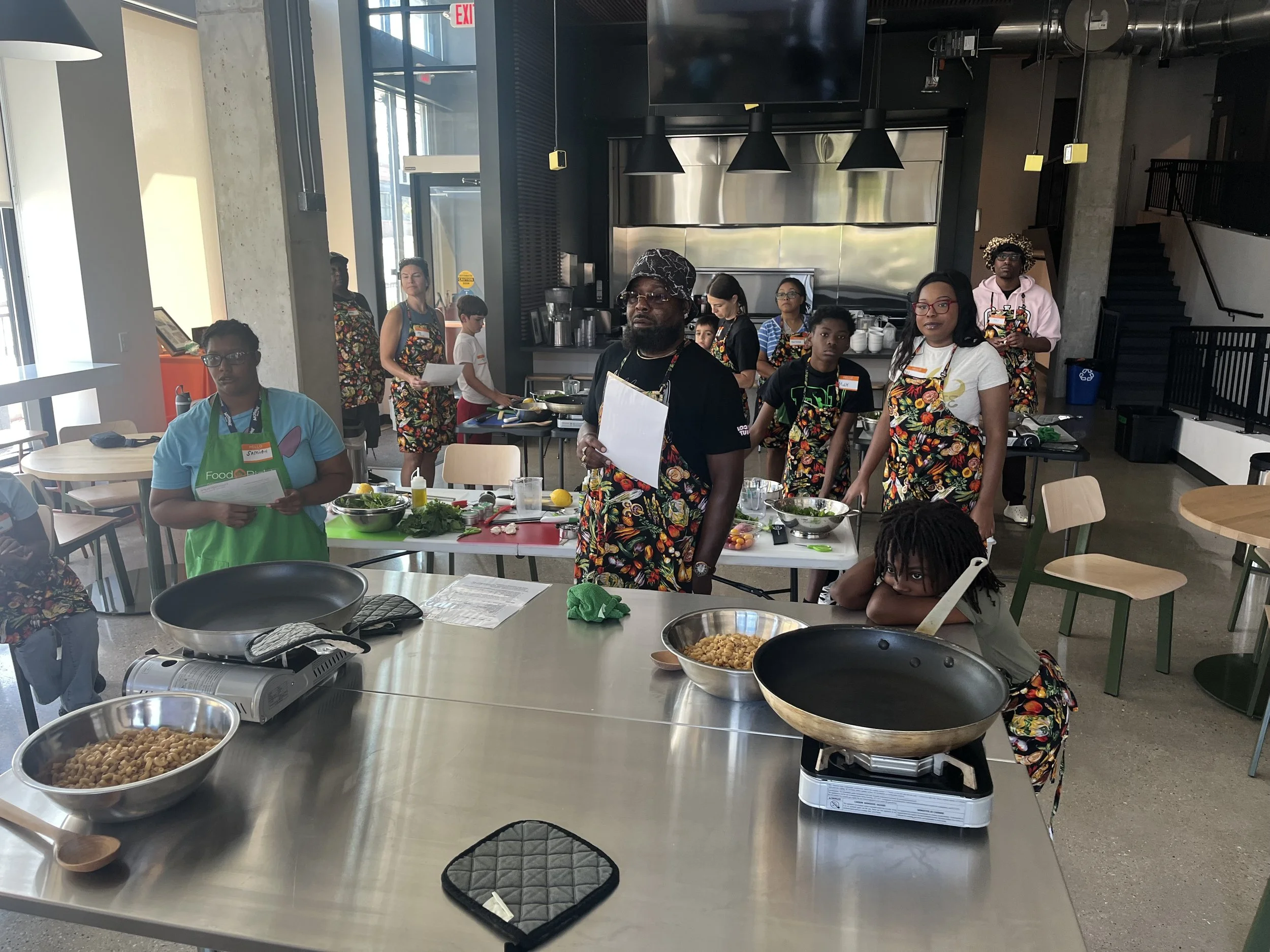 Group of people in a modern industrial-style kitchen participating in a cooking class or event, with ingredients and cooking utensils on the tables.