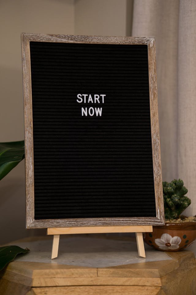 A black letter board with a wooden frame displaying the words "START NOW" in white letters, standing on a small easel on a wooden surface with plants nearby.