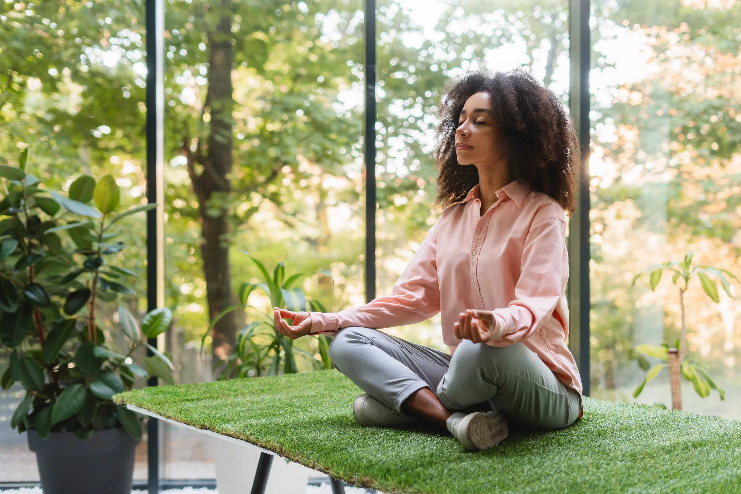 Une femme méditant en position de lotus dans une serre ou un espace intérieur, entourée de plantes et de grandes fenêtres avec vue sur un environnement extérieur verdoyant.