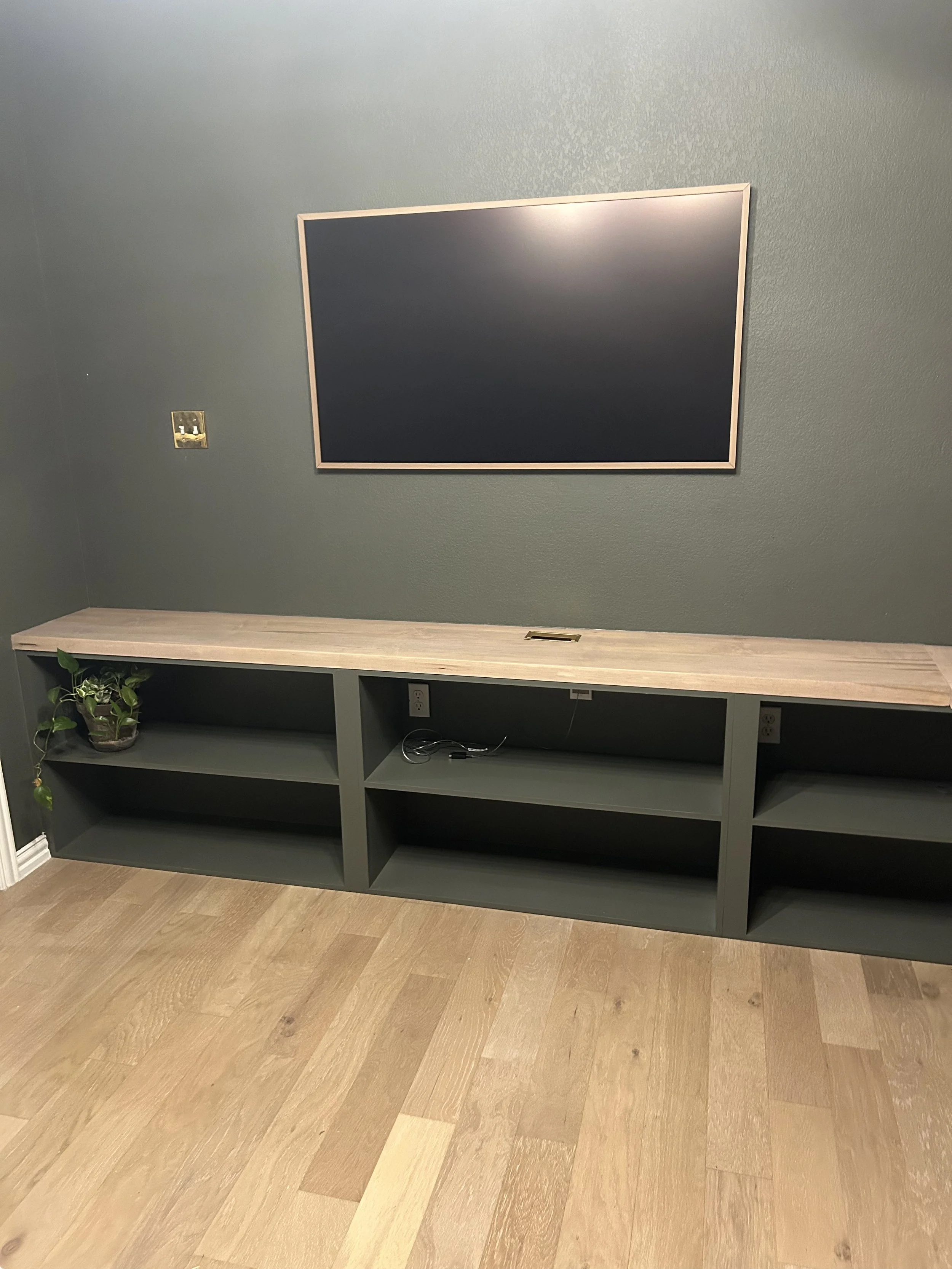 Living room with a large flat-screen TV mounted on the wall above a green entertainment unit with open shelves, a potted plant, and electrical outlets, with wooden flooring.