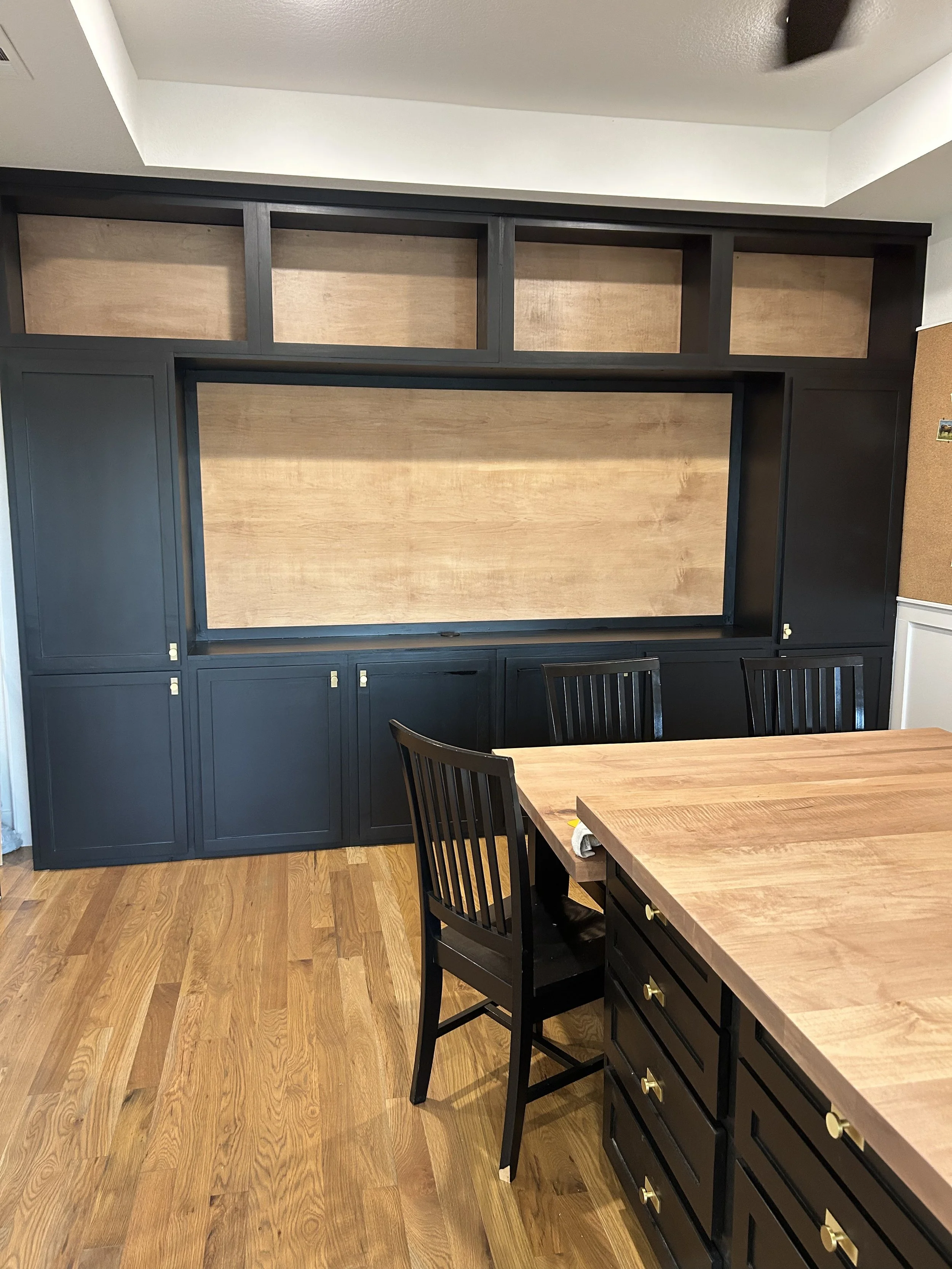 A large wooden entertainment center with black and natural wood finish in a kitchen or dining room, with a wooden table and black chairs in the foreground.