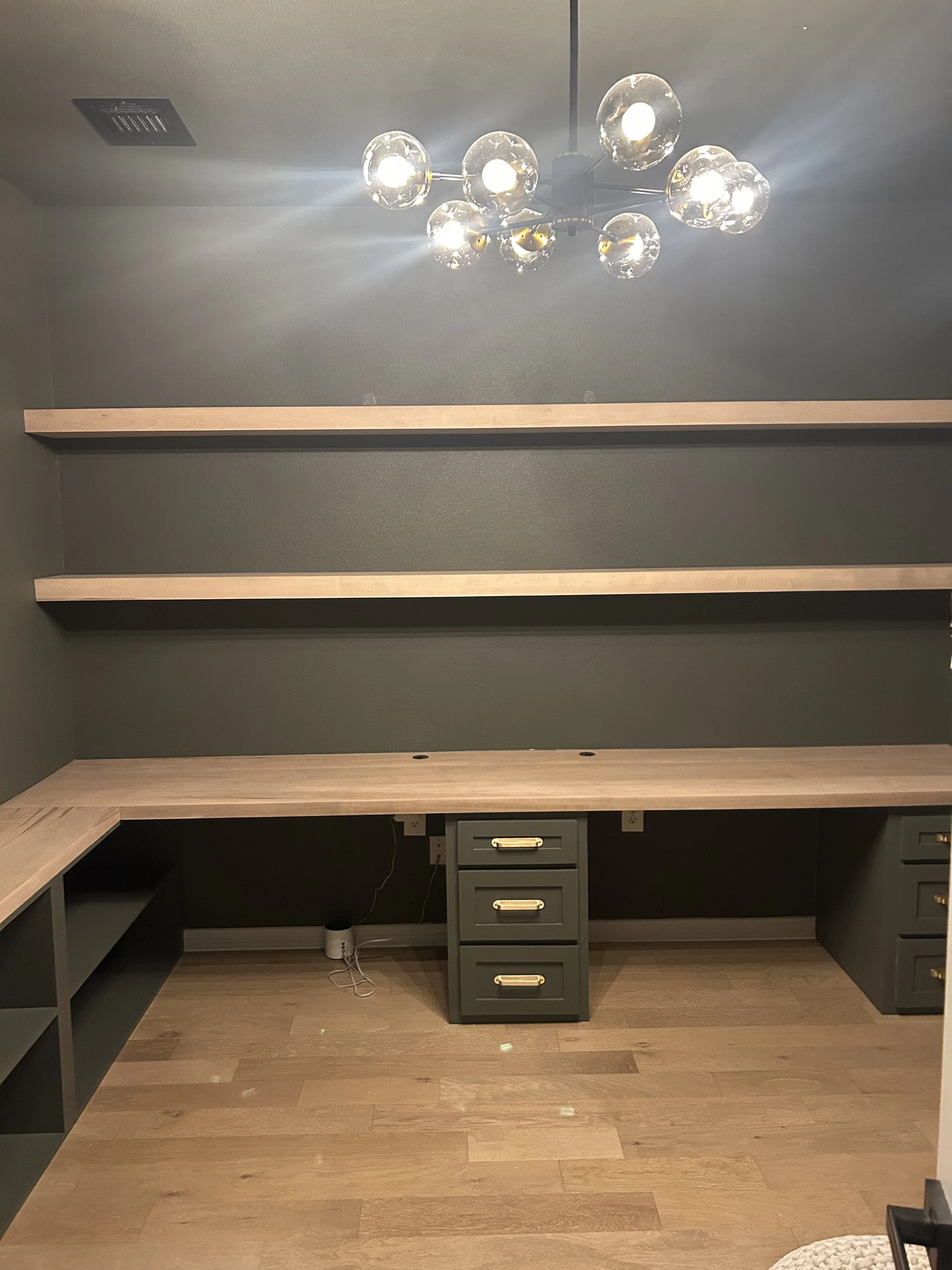 Empty home office with dark gray walls, a wooden desk, floating shelves, and a black filing cabinet, illuminated by a modern chandelier.