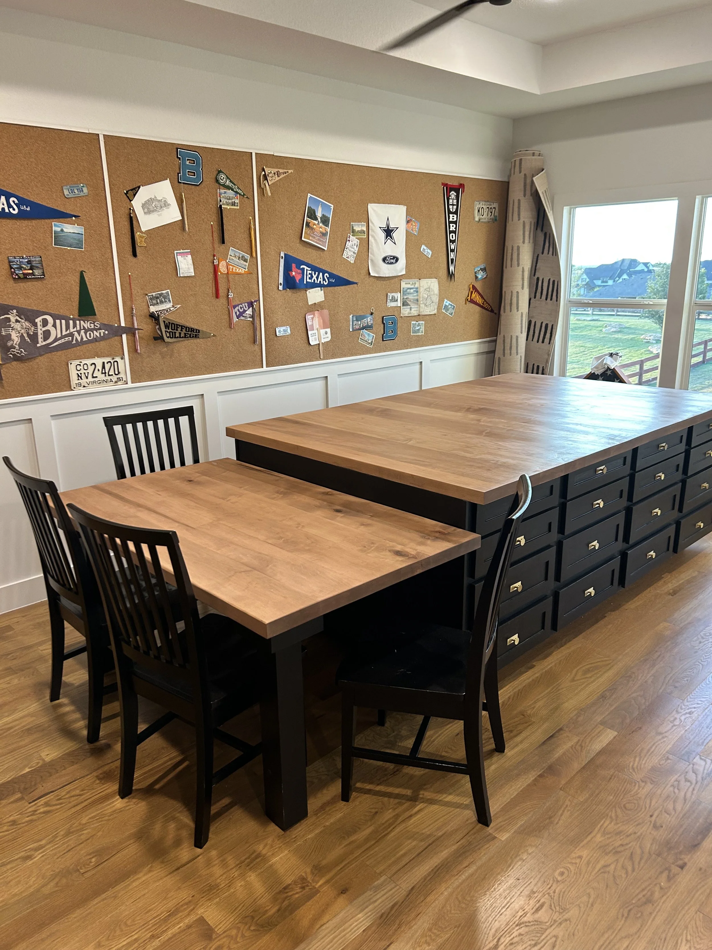 A spacious room with a large wooden table, four black chairs, and a cork bulletin board decorated with sports team pennants and photos, with a window showing a green outdoor landscape.