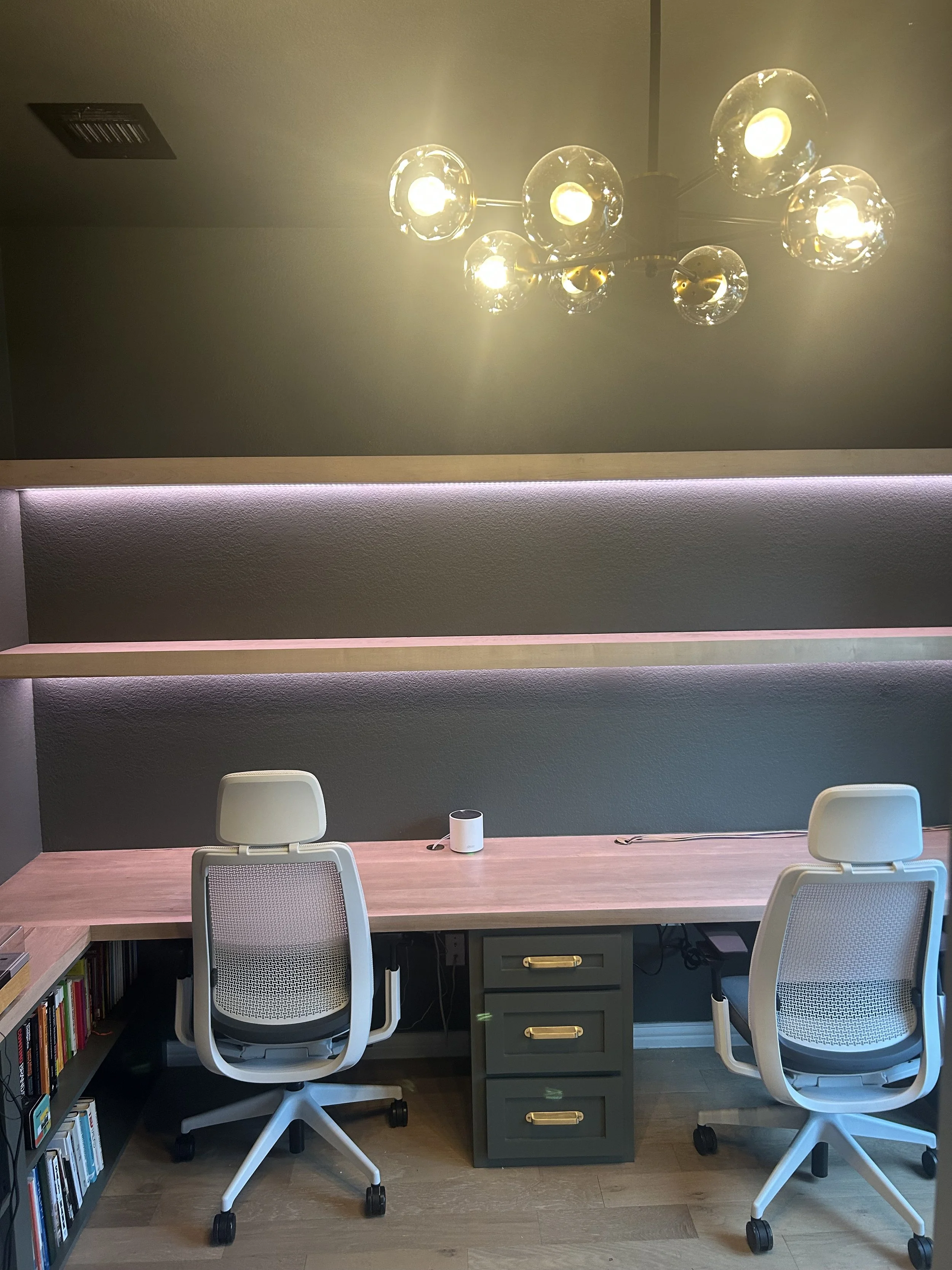 A modern home office with a pink wood desk, two white ergonomic office chairs, a gray three-drawer filing cabinet, a bookshelf, and a chandelier with multiple exposed bulbs hanging from the ceiling.