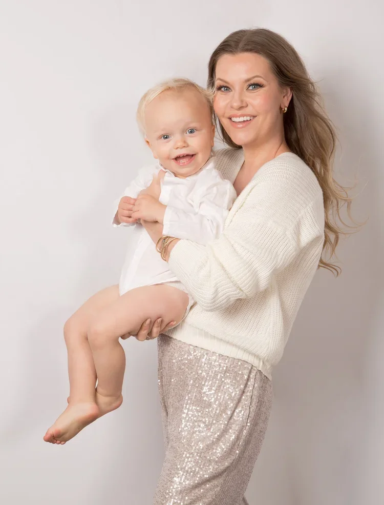 A woman with long brown hair holding a smiling blonde toddler girl, both looking at the camera, against a plain white background.