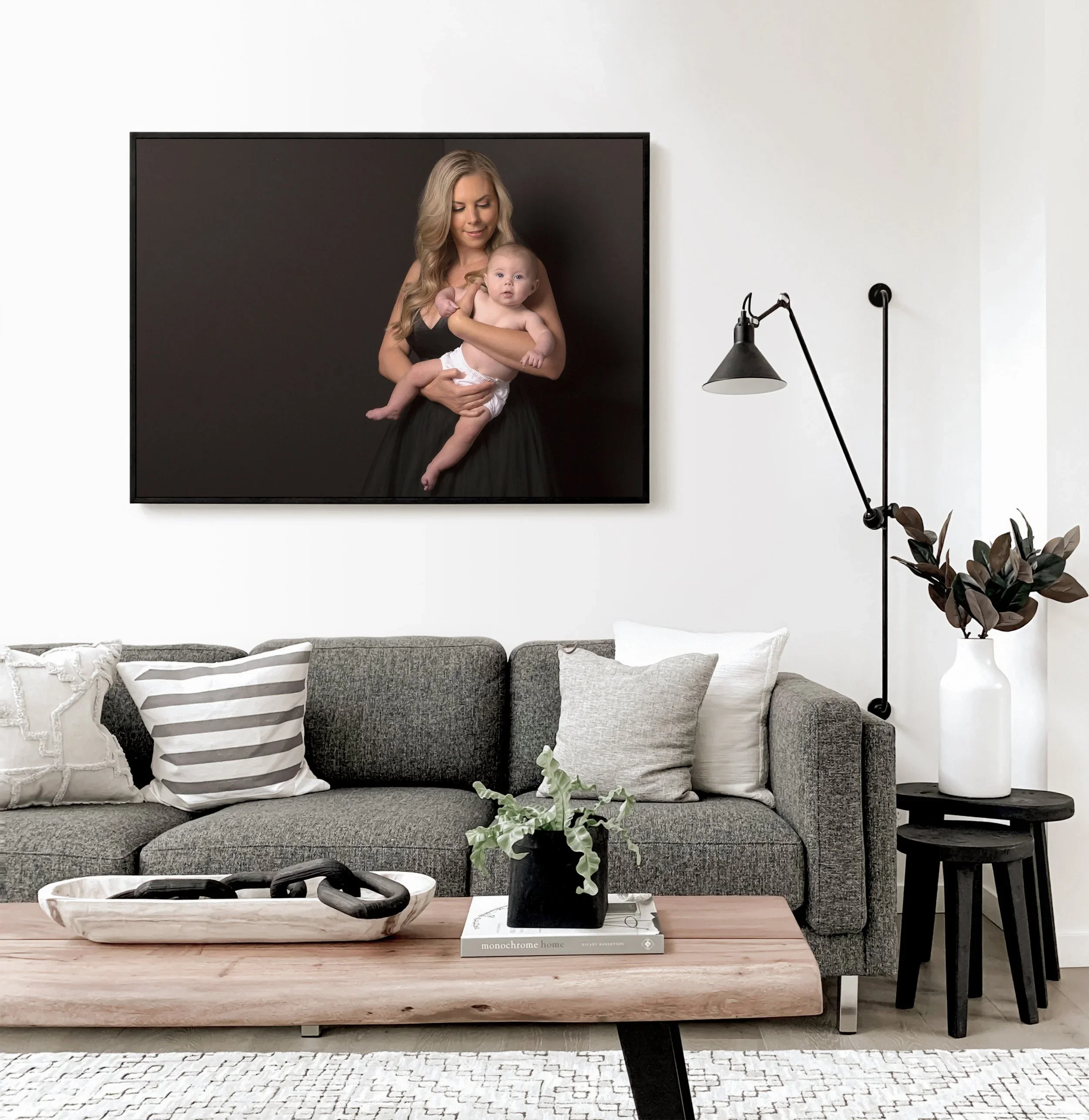 Living room with gray sofa, decorative pillows, wooden coffee table with plant and sculpture, black side table, white vase with dark leaves, wall-mounted black lamp, framed photo of a woman holding a baby