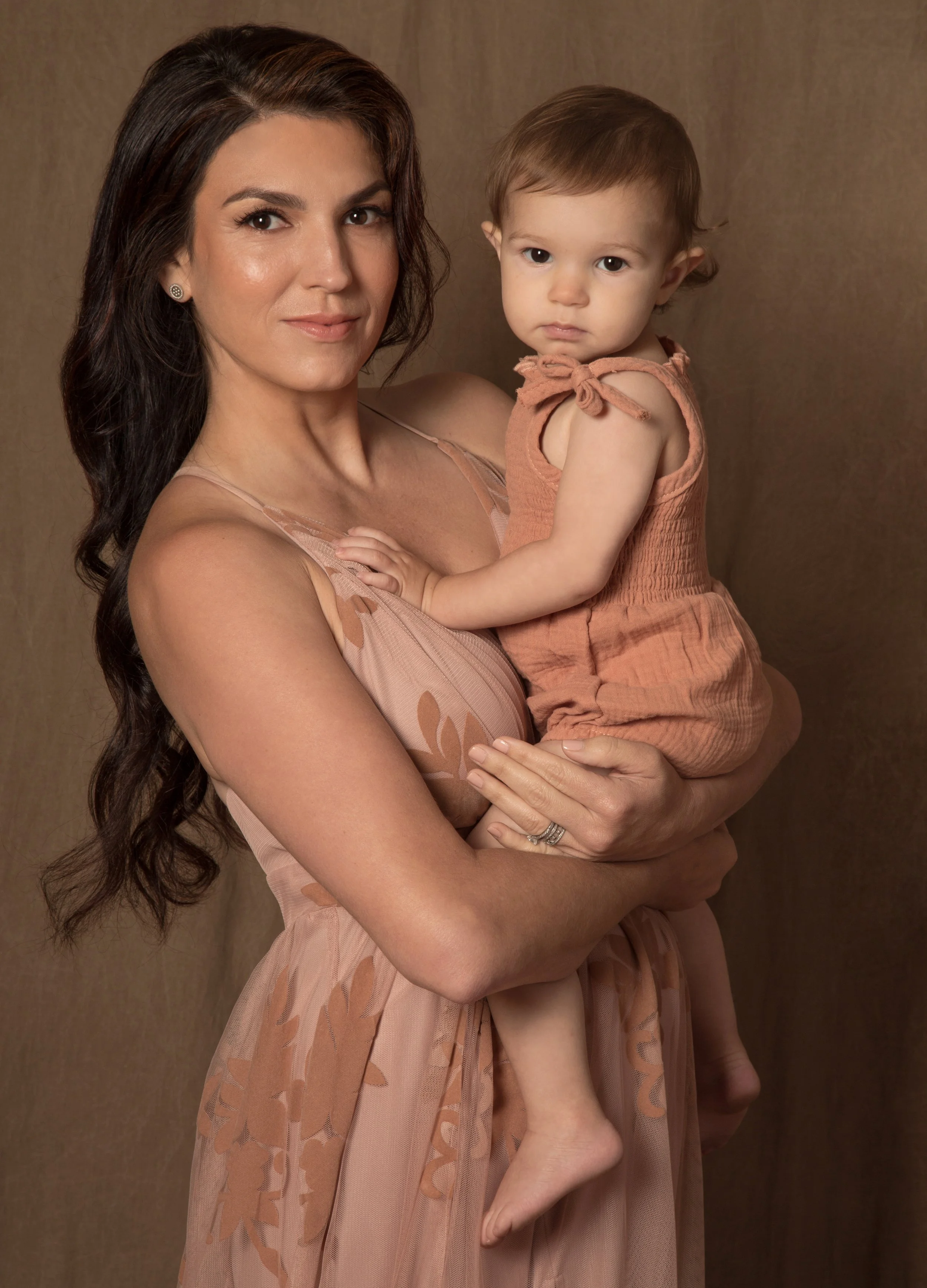 A woman with long, wavy dark brown hair holding a young girl with light brown hair and fair skin, both looking at the camera against a plain brown background.