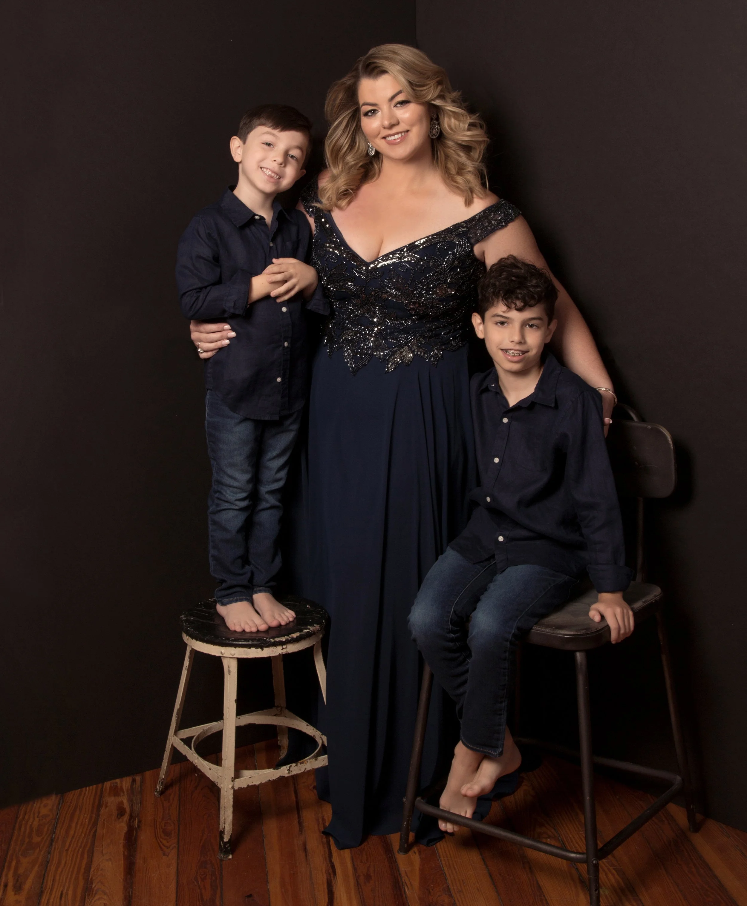 A woman dressed in a dark blue evening gown with two boys in matching dark blue shirts, posing together against a dark background. One boy stands on a small stool, and the other sits on a tall chair, all smiling.