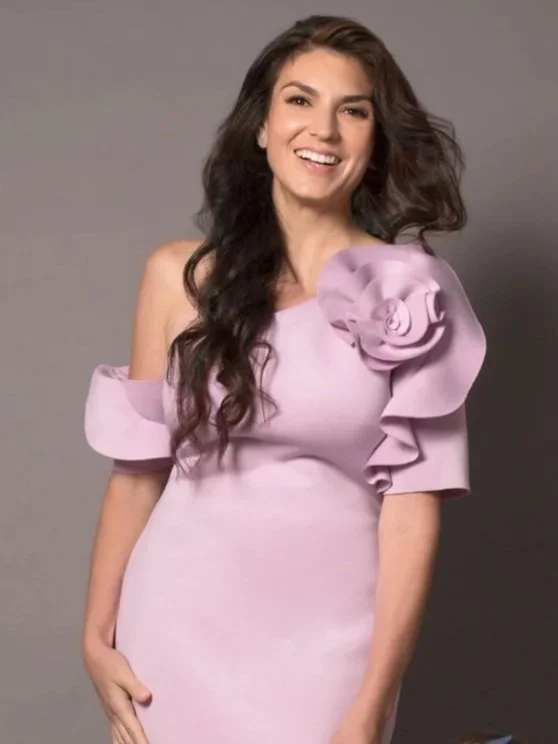 A woman with long dark hair wearing a light lavender dress with large ruffled sleeves and a decorative floral detail on one shoulder, smiling against a plain gray background.
