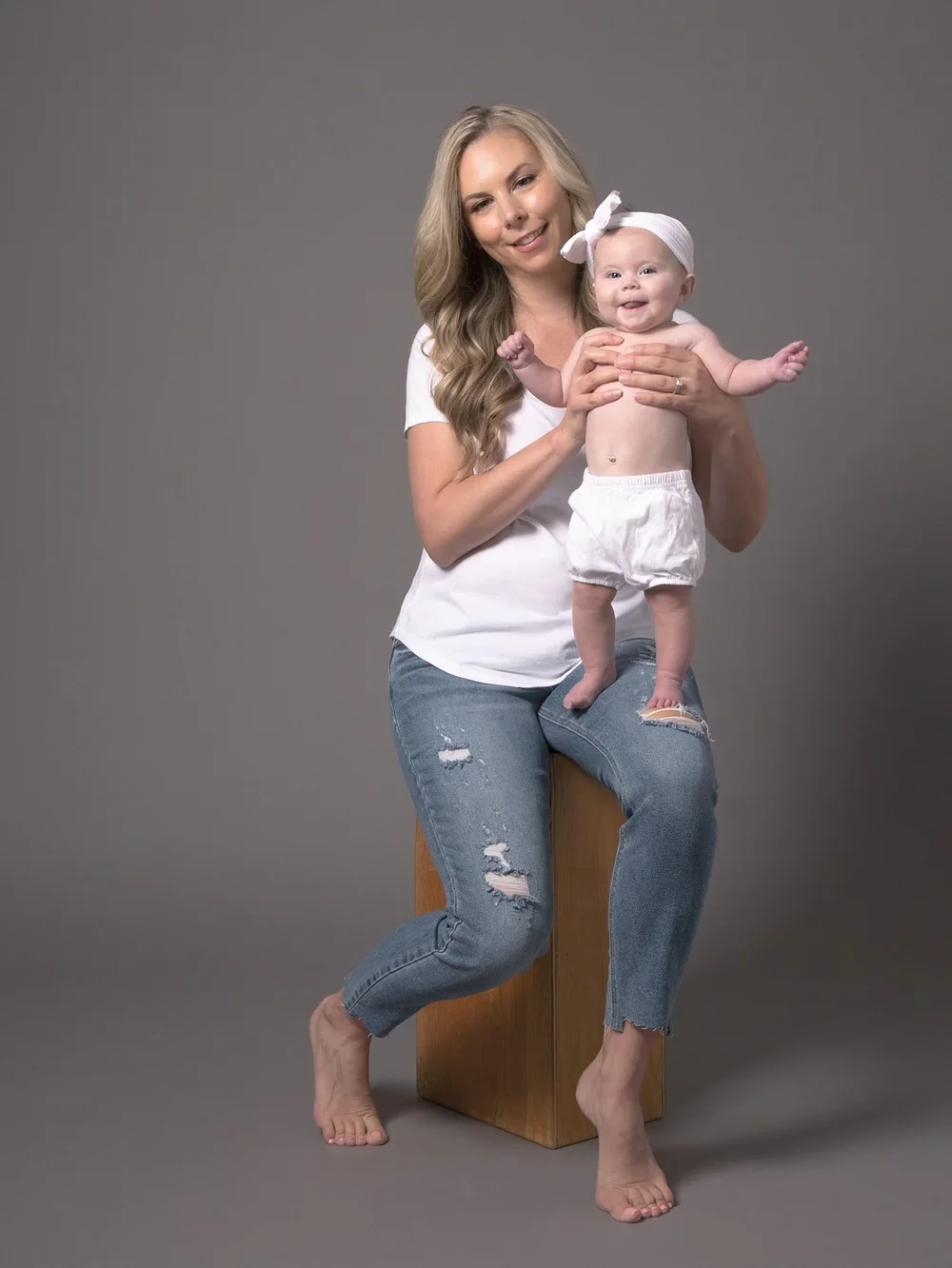 A woman with long blonde hair and wearing a white t-shirt and ripped jeans is sitting on a wooden block, holding a baby girl dressed in a white headband and diaper, against a gray background.