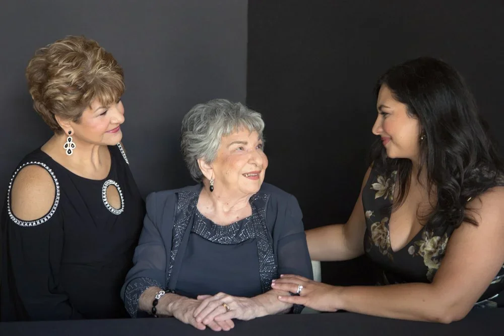 Three women, one elderly and two middle-aged, sitting and engaging in a friendly conversation in front of a plain dark backdrop.