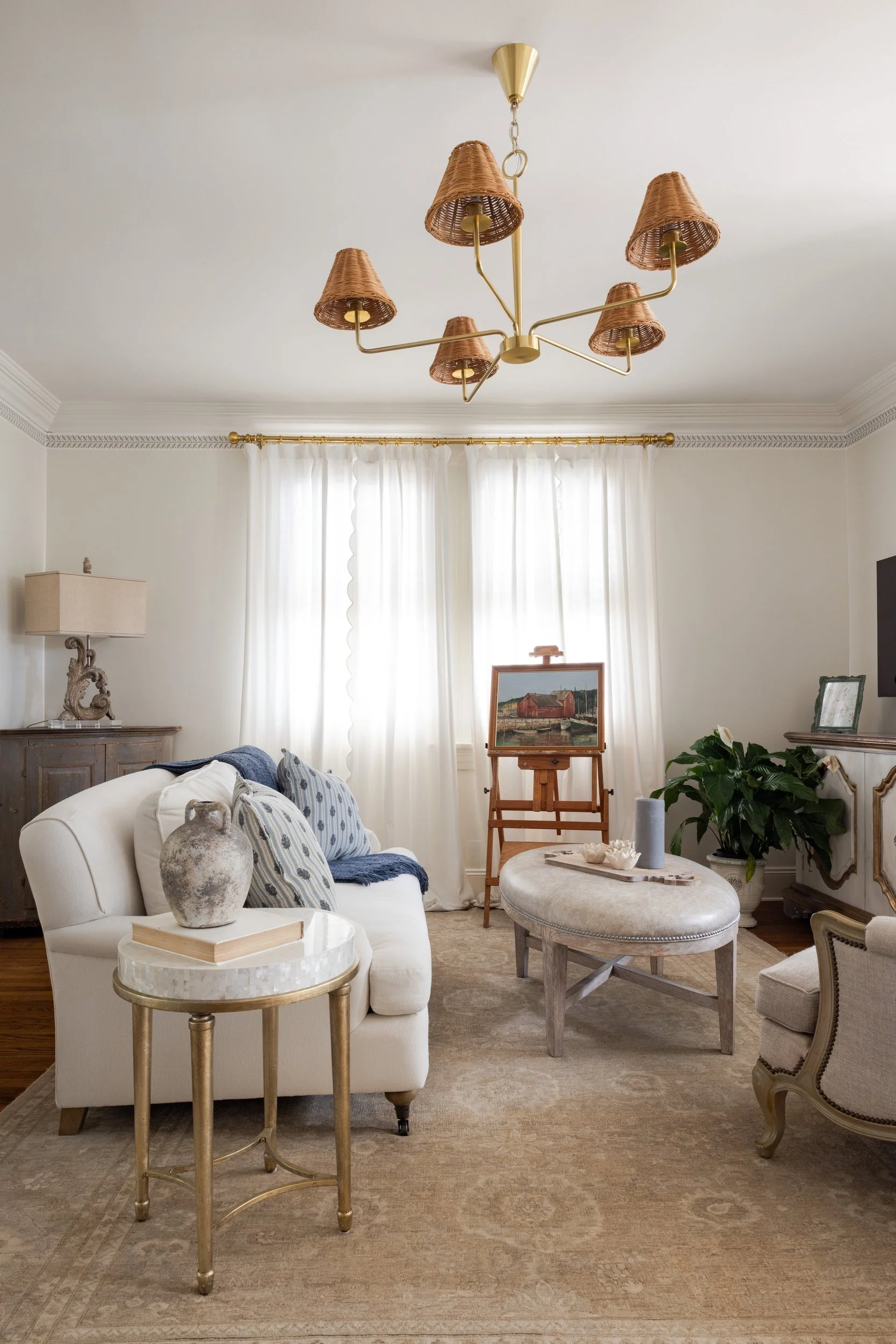 Living room with a brass and wicker chandelier, scalloped white curtains, and linen sofa