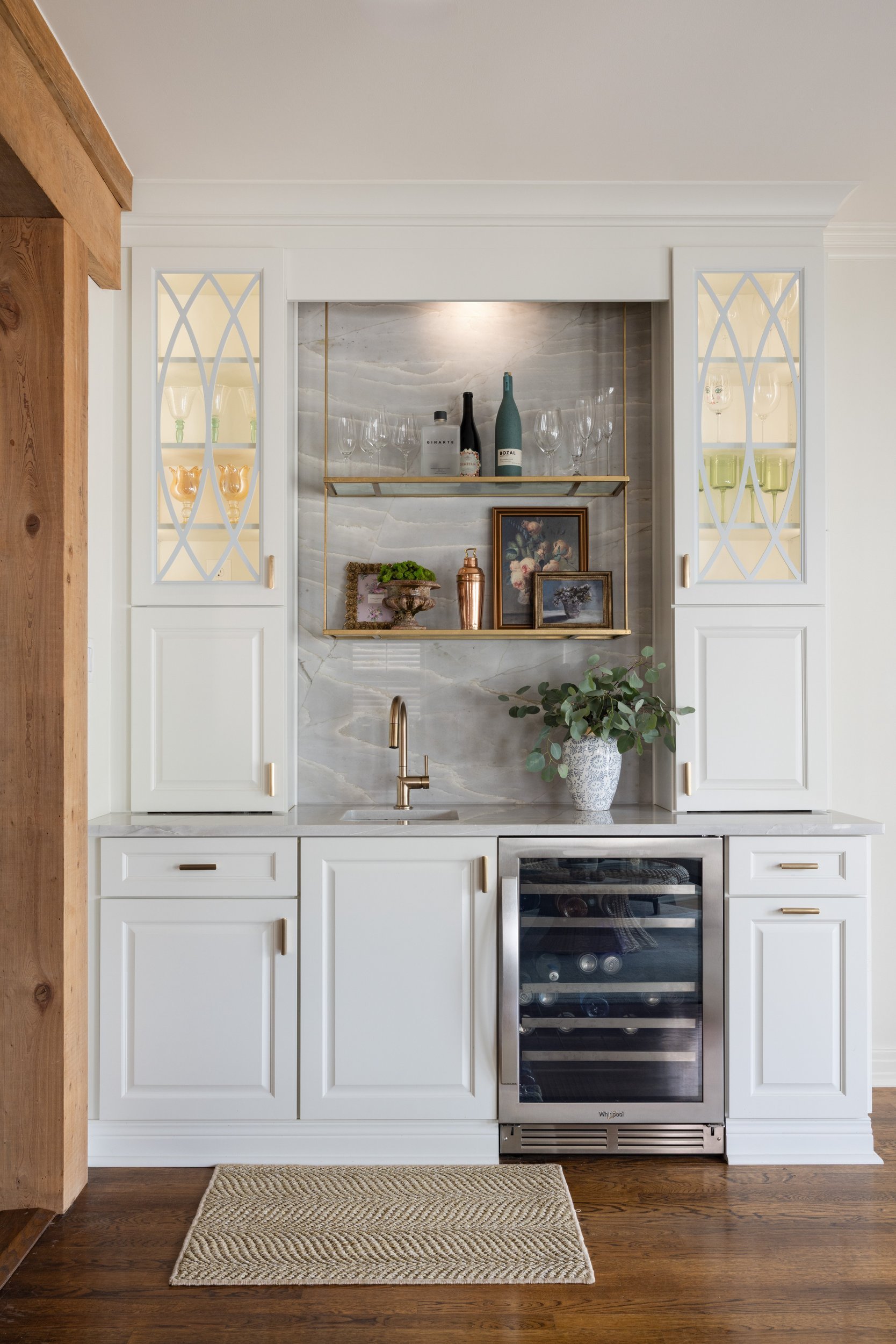 Built-in bar with lit glass cabinets, brass shelves, quartzite backsplash, and wine fridge