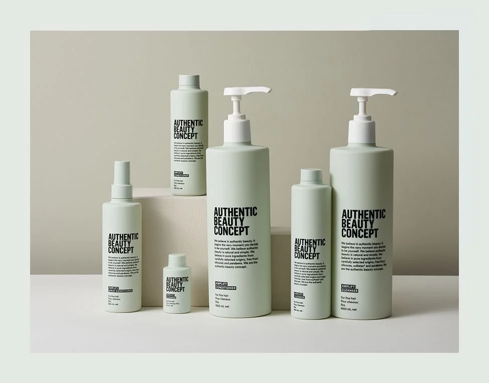Collection de produits cosmétiques de la marque Authentic Beauty Concept, comprenant des bouteilles de shampoing, après-shampoing, conditionneur et nettoyant, de différentes tailles et formes, sur un support blanc et devant un fond beige.