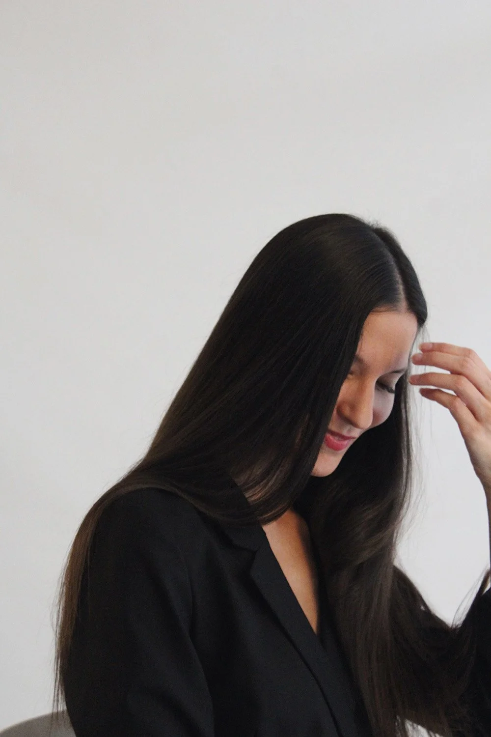 Femme aux cheveux longs et lisses, portant une chemise noire, levant la main près de son visage, contre un mur blanc.