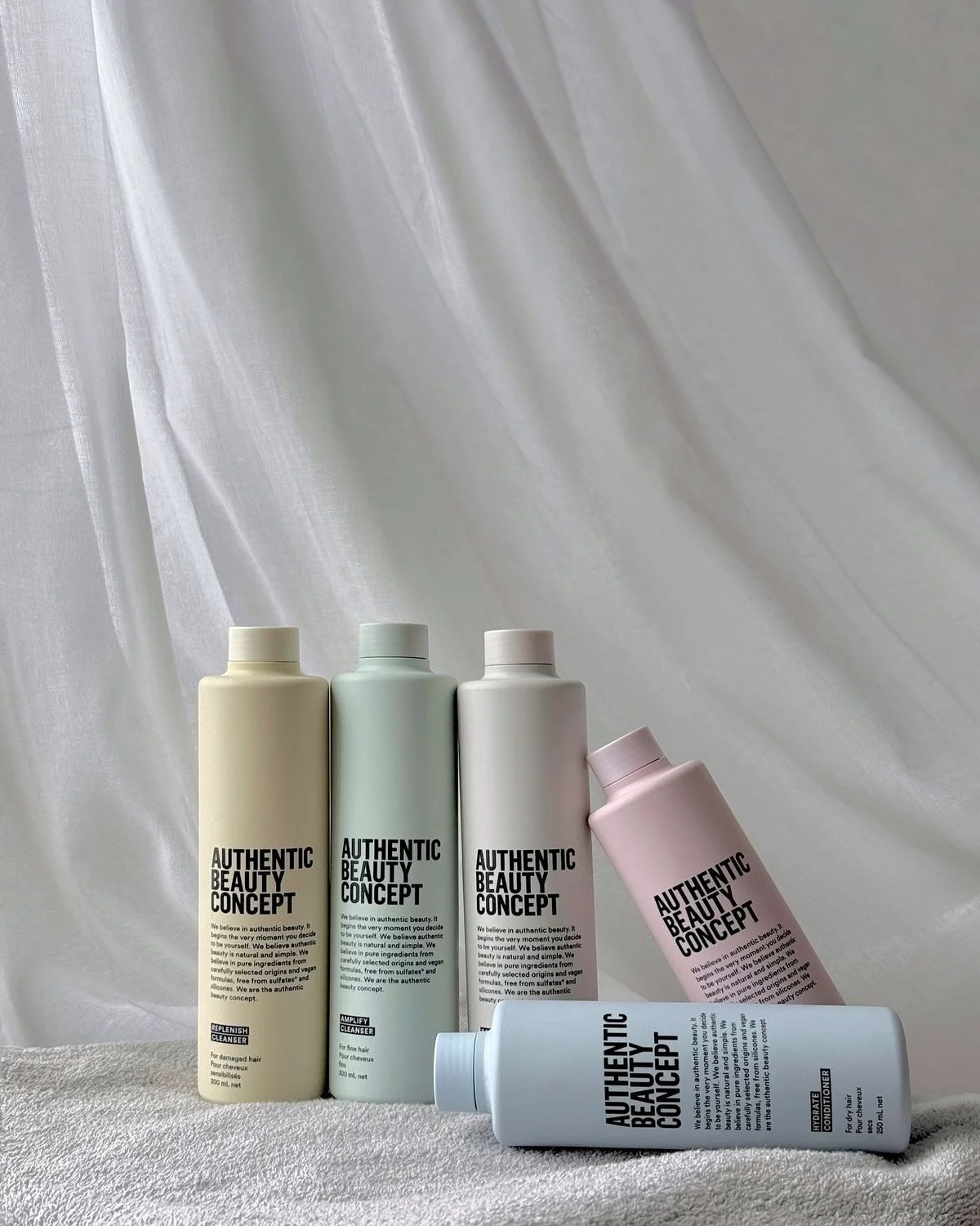 Cinq bouteilles de produits de soins pour cheveux de la marque authentic beauty concept, avec différentes couleurs, posées sur une surface douce avec un fond en tissu blanc léger.