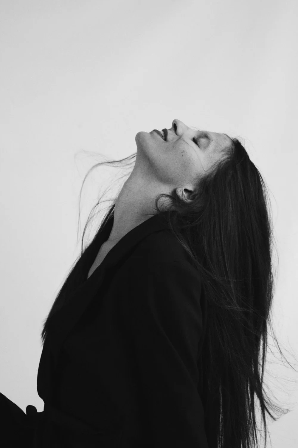 Portrait en noir et blanc d'une femme à l'air joyeux, les yeux fermés, la tête inclinée en arrière, cheveux longs et sombres, portant un vêtement noir