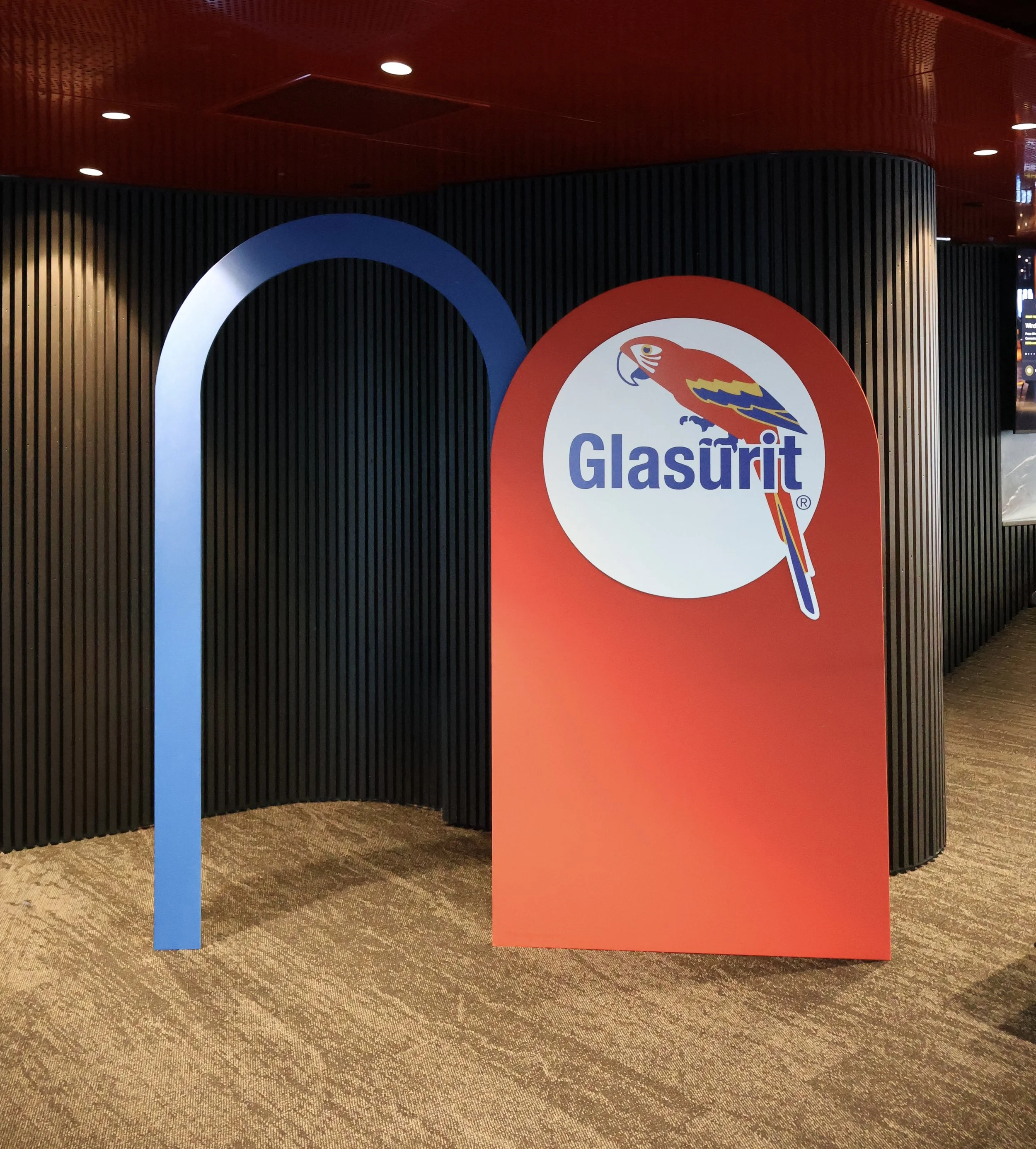 Sign with the Glascurit logo, featuring a stylized colorful parrot and a blue letter 'Q' in the background, situated on a red display board in a modern indoor space.