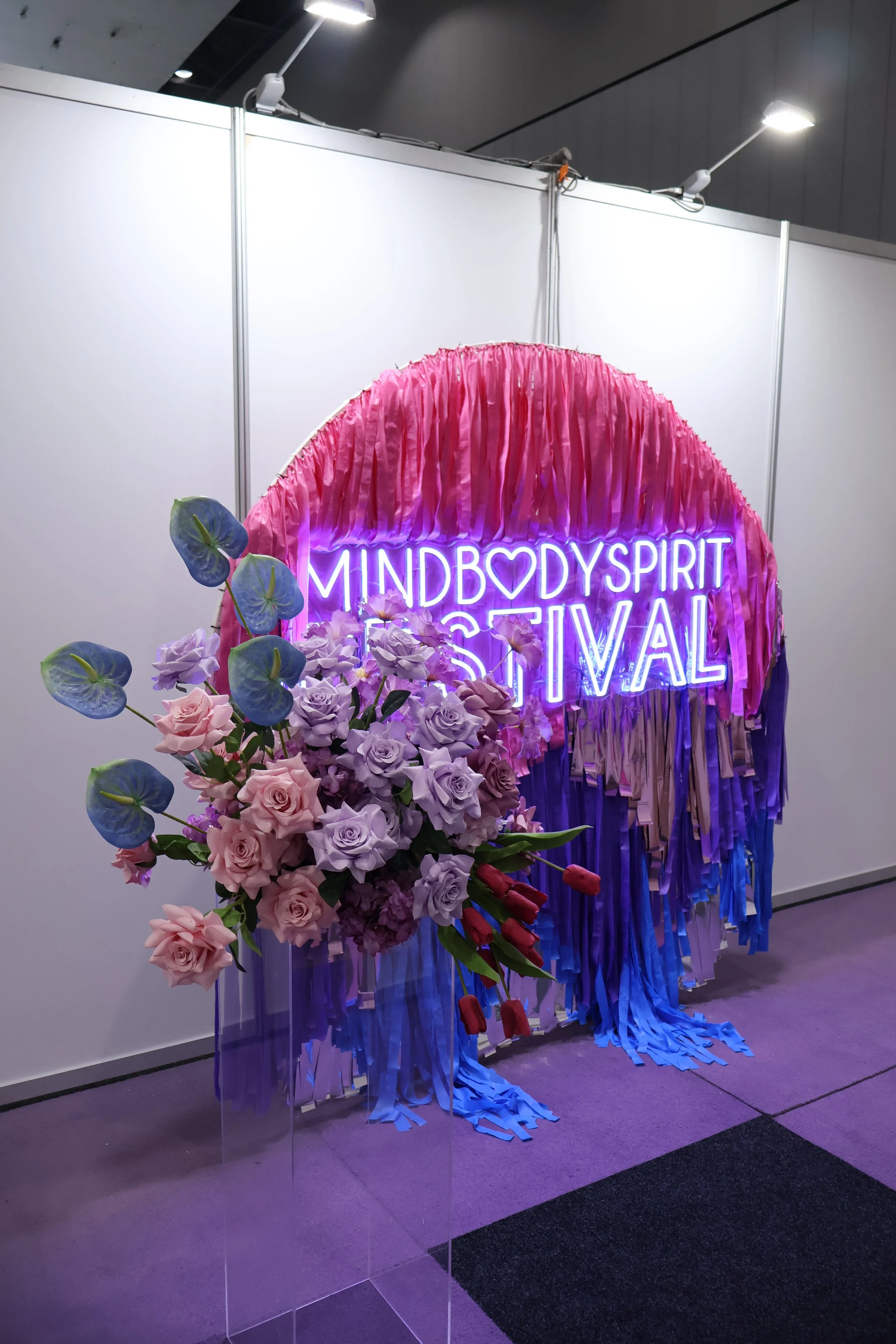 Decorative installation with a pink fringe backdrop, neon sign saying 'Mind Body Spirit Festival', and a bouquet of flowers including purple roses, white roses, and blue anthuriums. The installation is in an indoor space with white walls and overhead lighting.