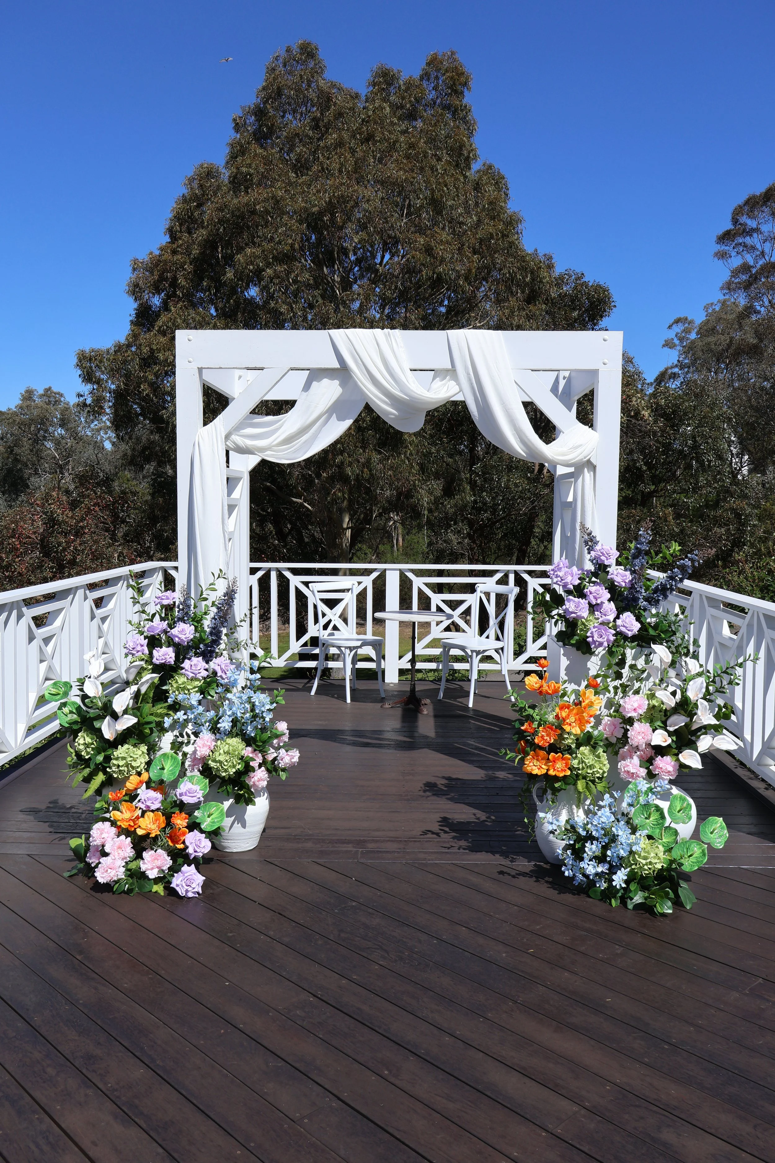 A white wedding arch with draped fabric on a wooden deck, decorated with potted flowers, against a backdrop of tall trees and a clear blue sky.