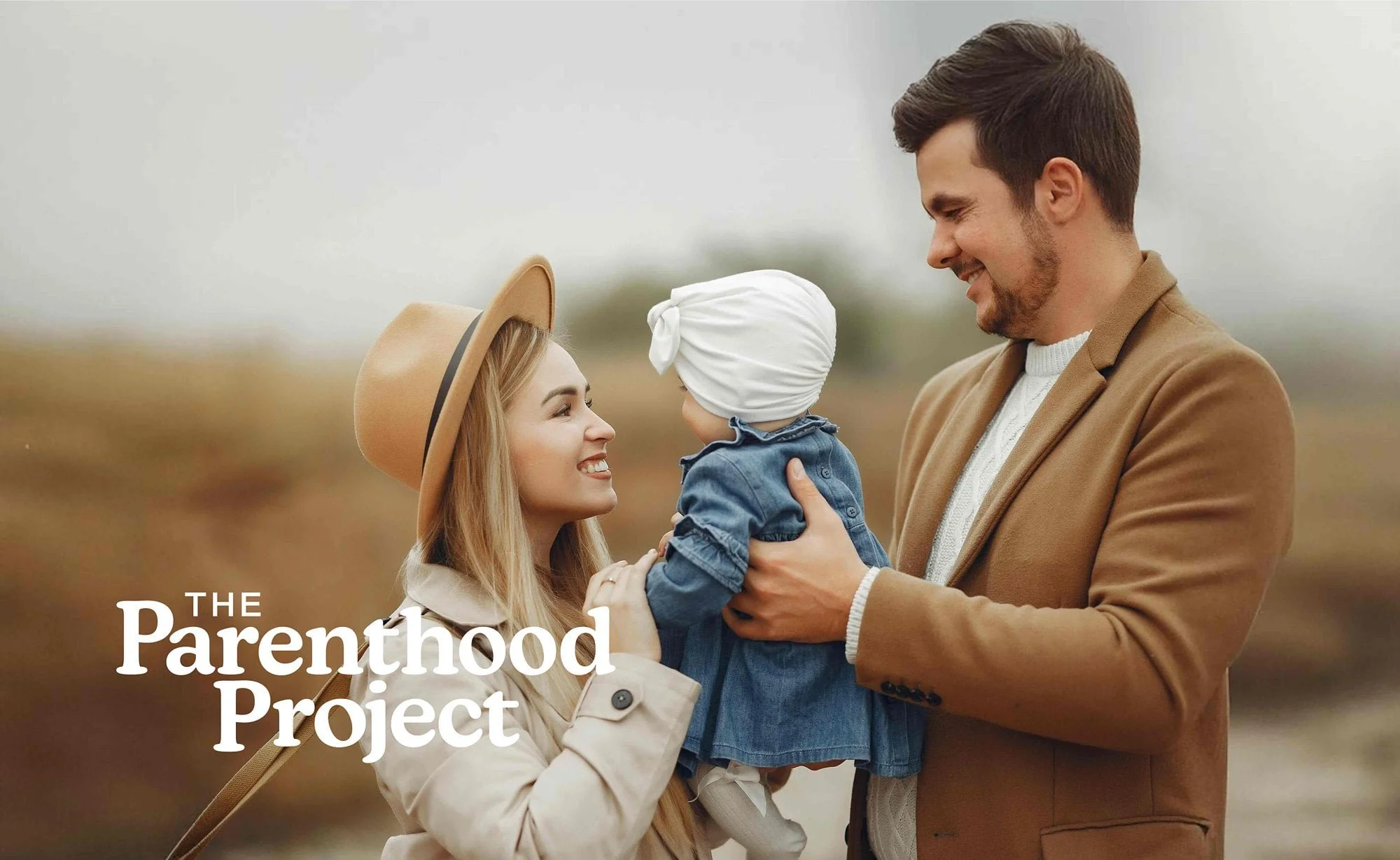 A young couple with a small child outdoors in a field, smiling at each other. The woman is wearing a tan wide-brimmed hat and a beige coat, the man is in a brown blazer, and the child is in a denim dress with a white head covering. Text overlay says 'The Parenthood Project'.