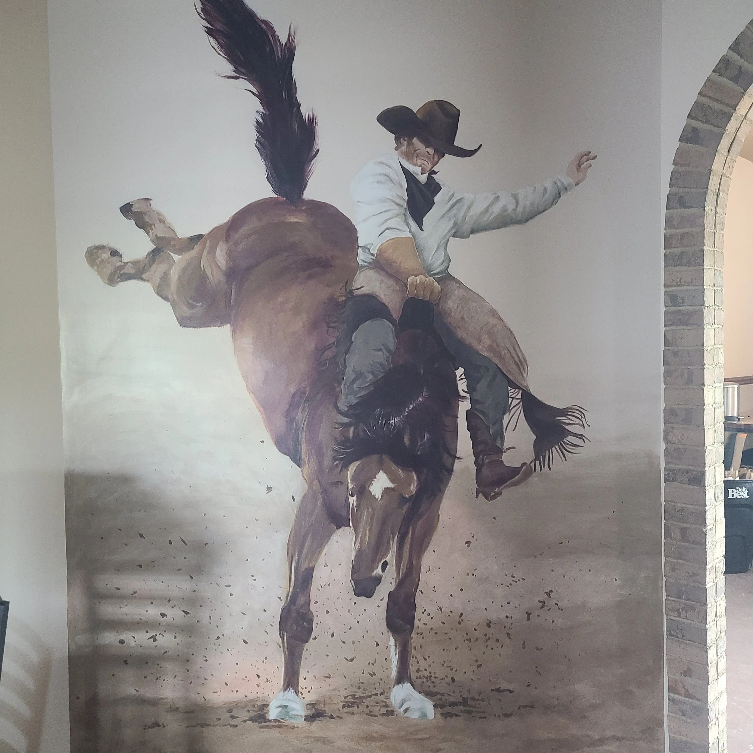 A mural of a cowboy riding a bucking horse, wearing a wide-brimmed hat and white shirt, on a wall next to a brick archway.