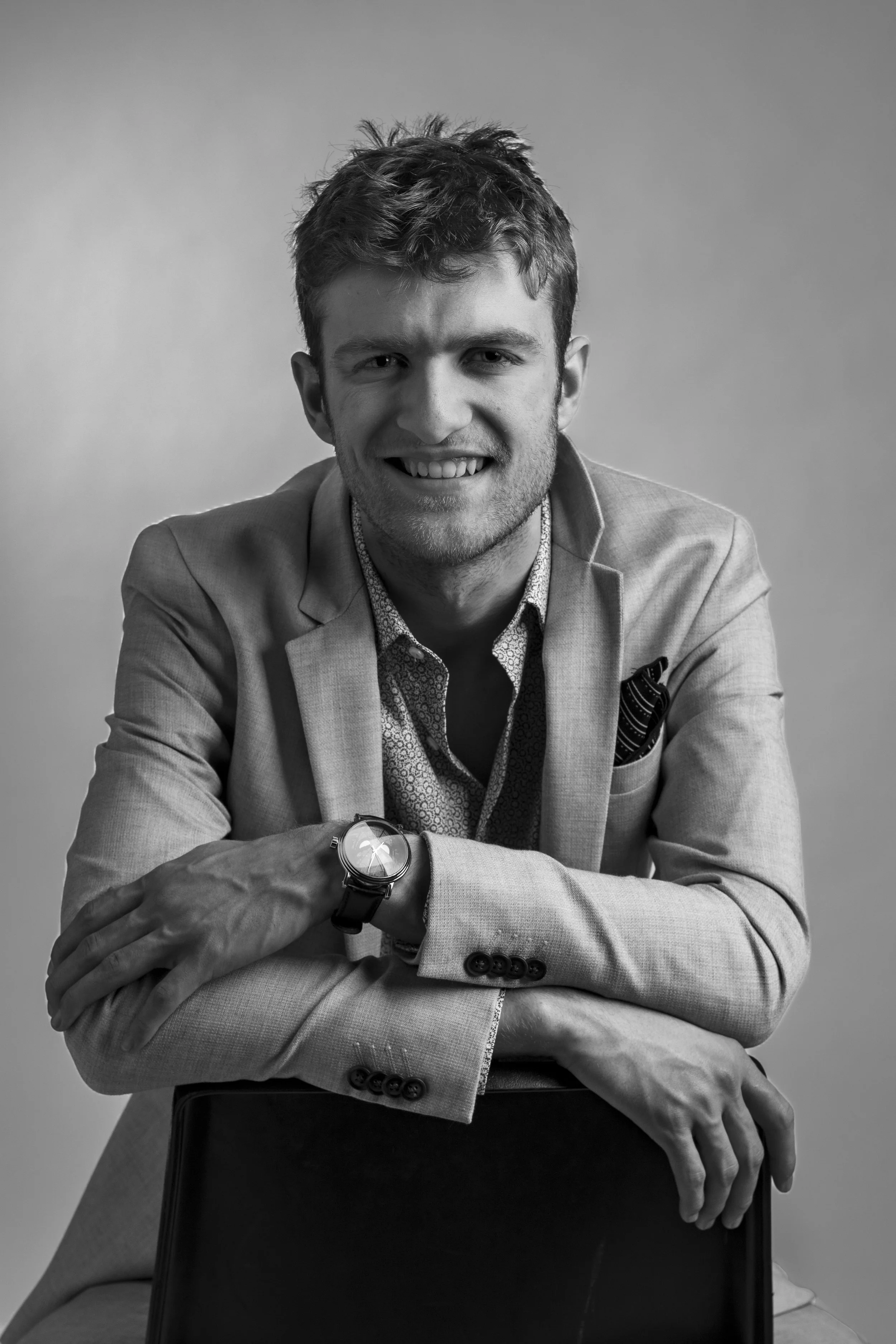 Black and white portrait of a young man with light hair, smiling, wearing a suit jacket, patterned shirt, and wristwatch, sitting with arms crossed on a chair.