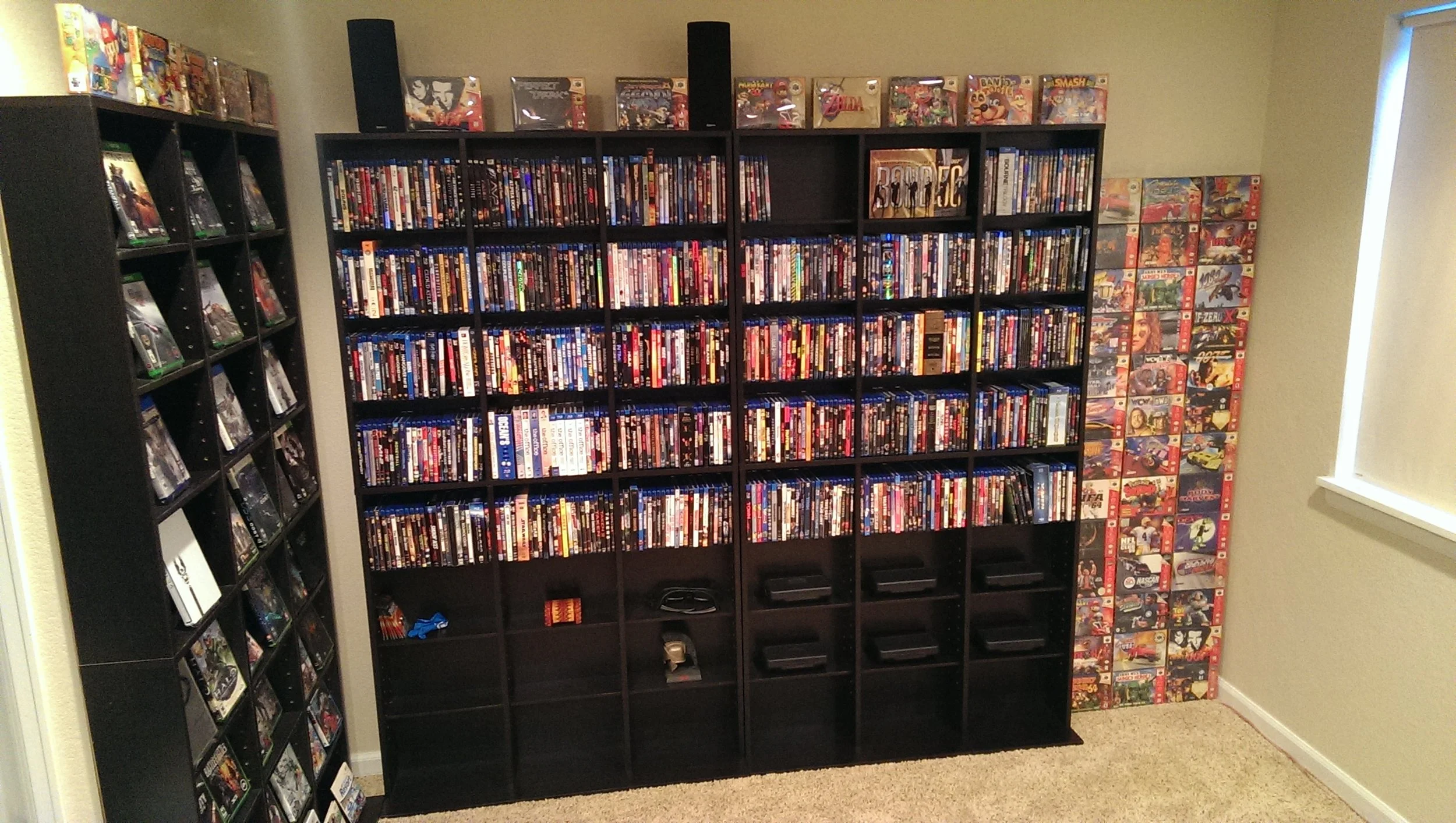 A room with black shelves filled with DVDs and video games, including a tall vertical shelf on the right side and a large black grid-style shelf in the center, topped with boxed video games or collectibles.