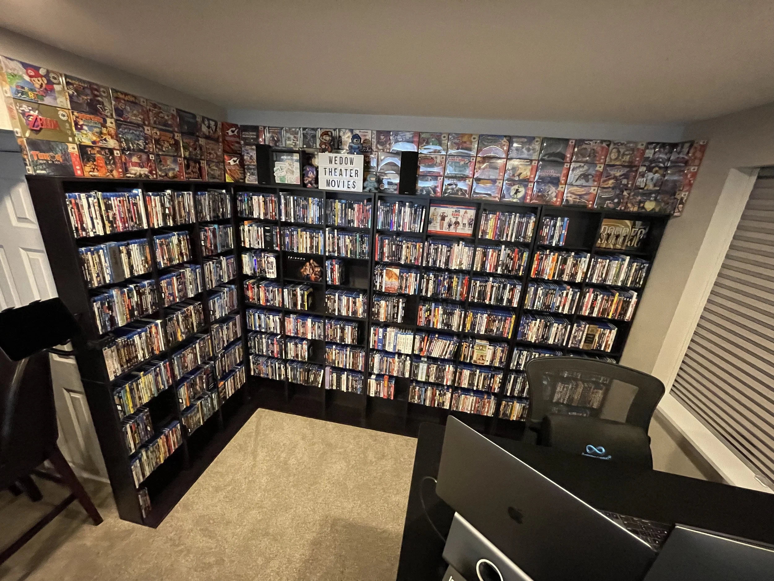 A room with black bookshelves filled with DVDs and movies, a sign that reads 'WEDOW THEATER MOVIES,' and a bookshelf with vintage video games on top.