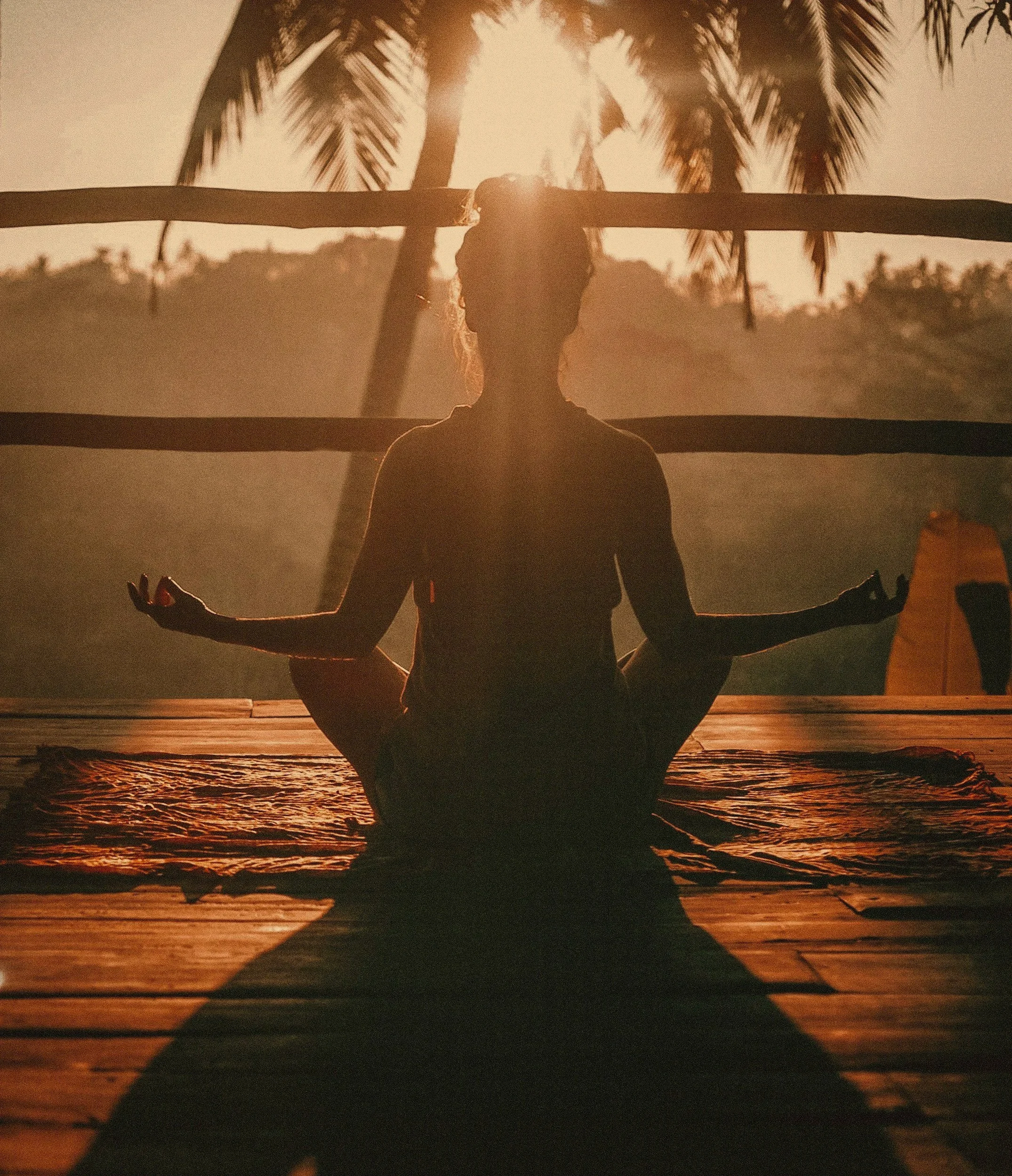 Person practicing yoga in a seated meditation pose on a wooden deck at sunset, with a backdrop of tropical palm trees and a calm ocean.