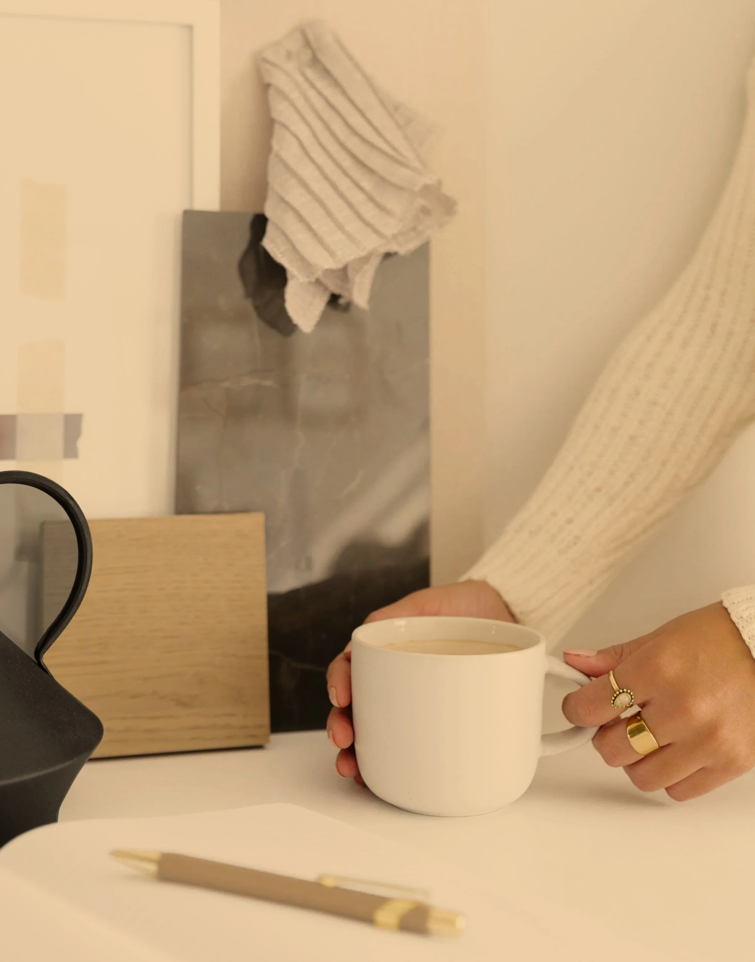 A woman holding a white coffee mug, wearing rings on her fingers and a beige long-sleeve top, sitting at a table with a notepad and golden pen, with a coffee machine in the background.