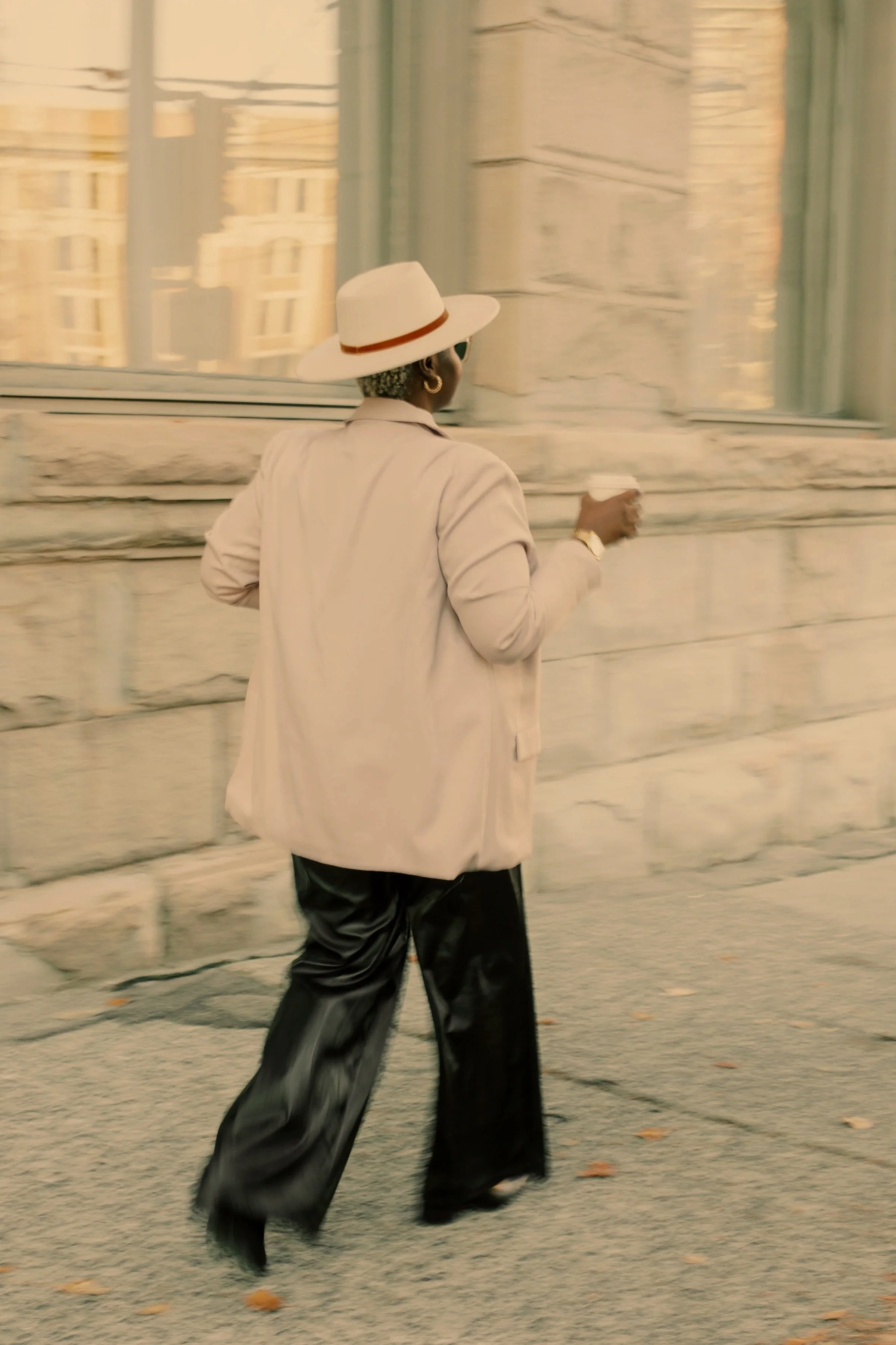 A person walking on a city sidewalk away from the camera, wearing a light-colored blazer, shiny black pants, a white wide-brimmed hat with a brown band, and sunglasses, holding a small cup in their right hand.