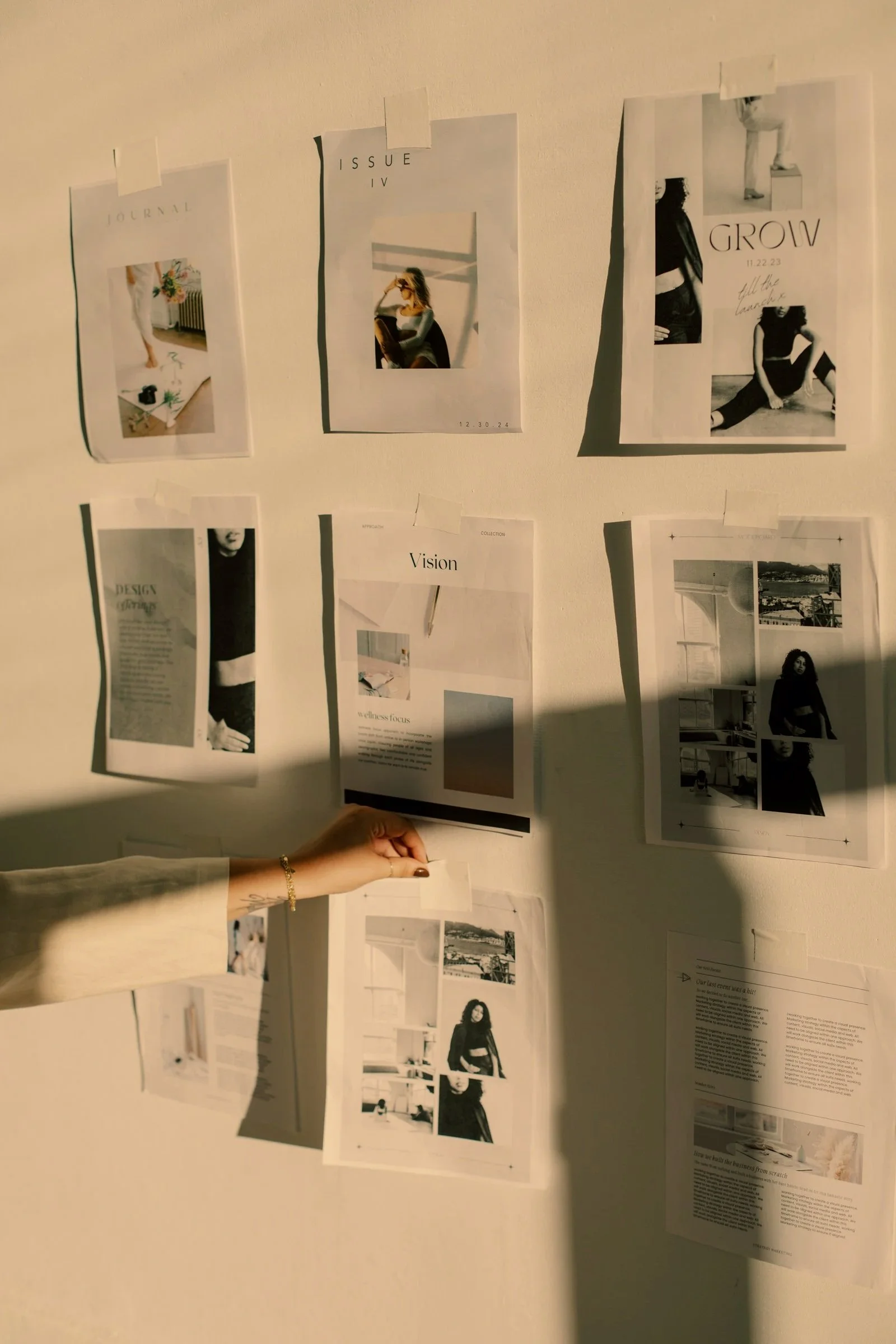 Wall with various printed pages and photographs taped to it, including images of women, text, and sketches. A person's hand is visible at the bottom holding a page.