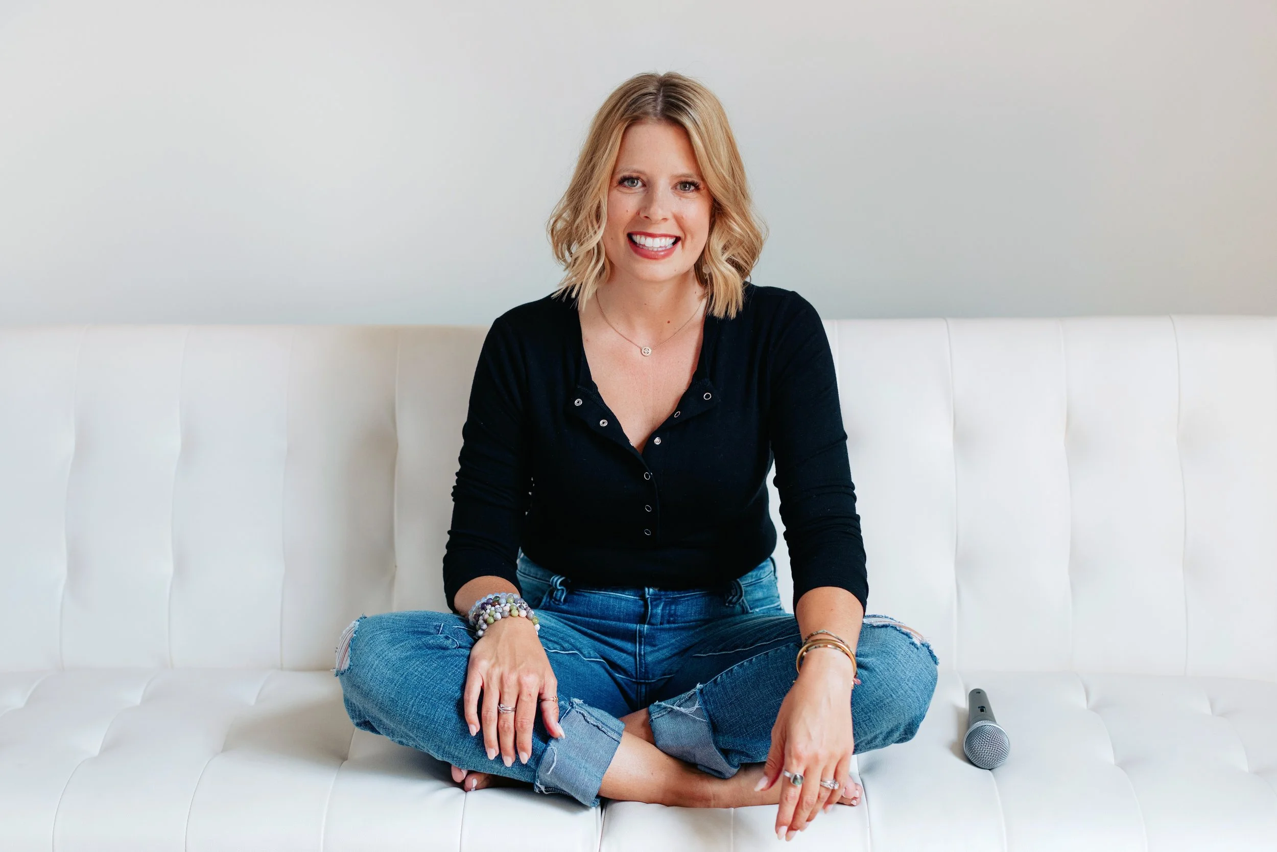A woman with blonde hair, smiling, sitting cross-legged on a white sofa, wearing a black top and ripped jeans, with colorful beaded bracelets on her wrist, a necklace, and a ring, with a microphone on the sofa beside her.