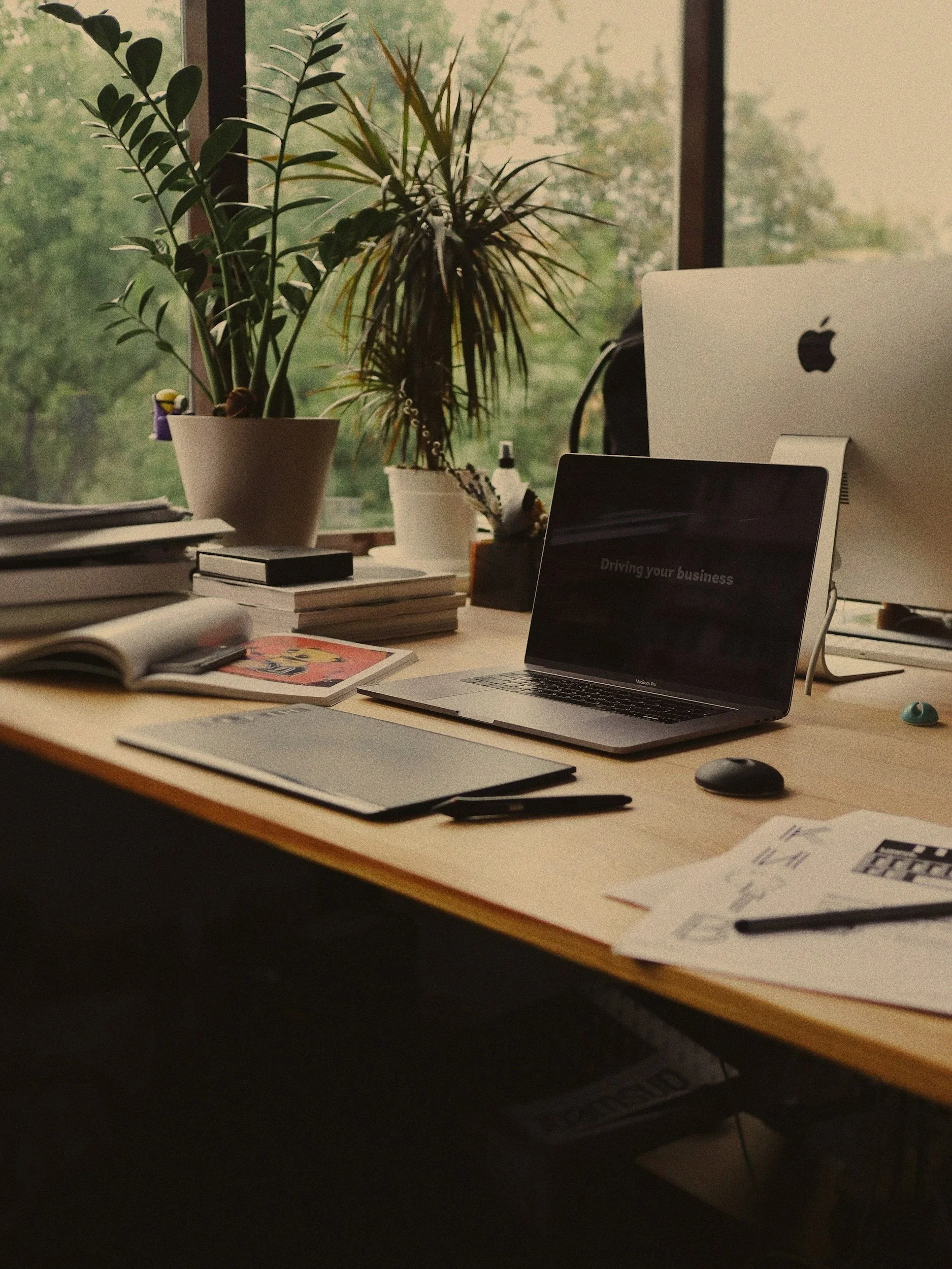 A cluttered desk with books, notebooks, a magazine, a pen, a mouse, a laptop, and an iMac monitor. There are two potted plants in the background near a window with green trees outside.
