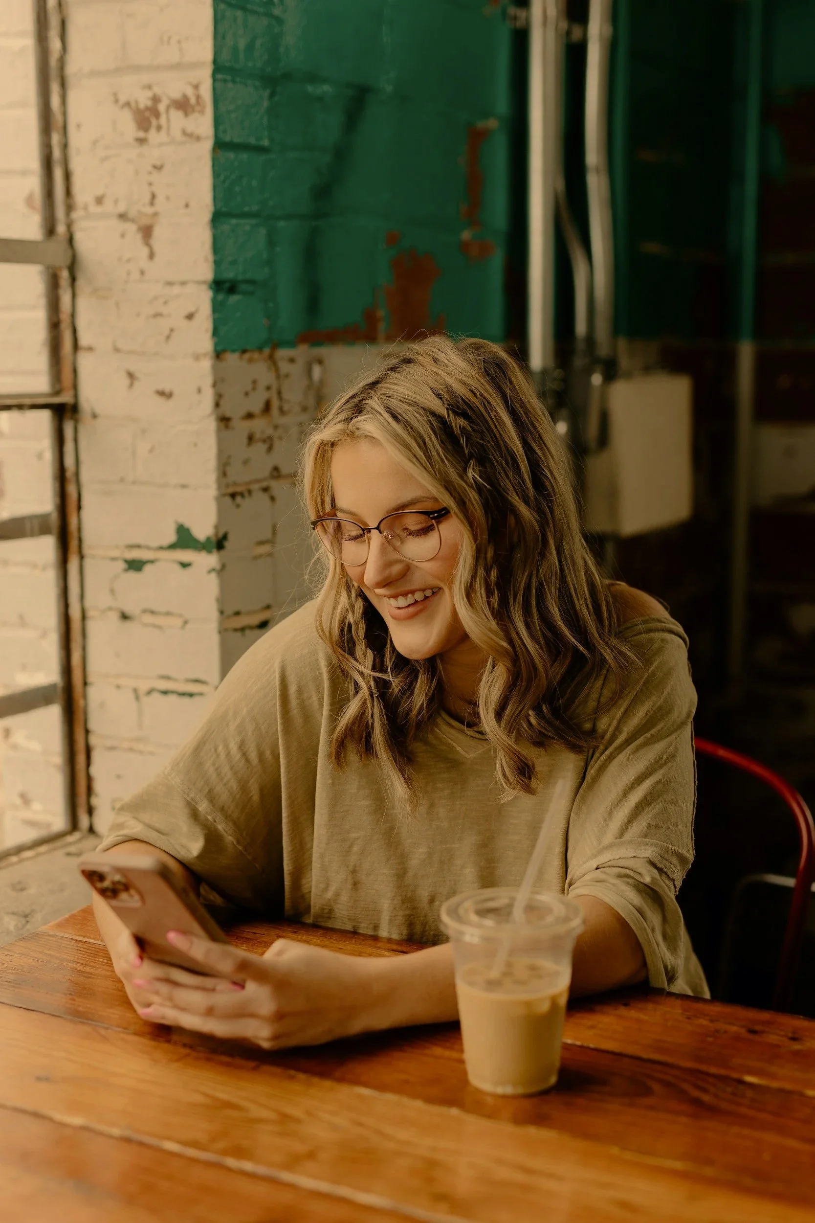 A young woman with glasses sitting at a wooden table, smiling while looking at her phone. There is a plastic cup with an iced beverage and a straw on the table.