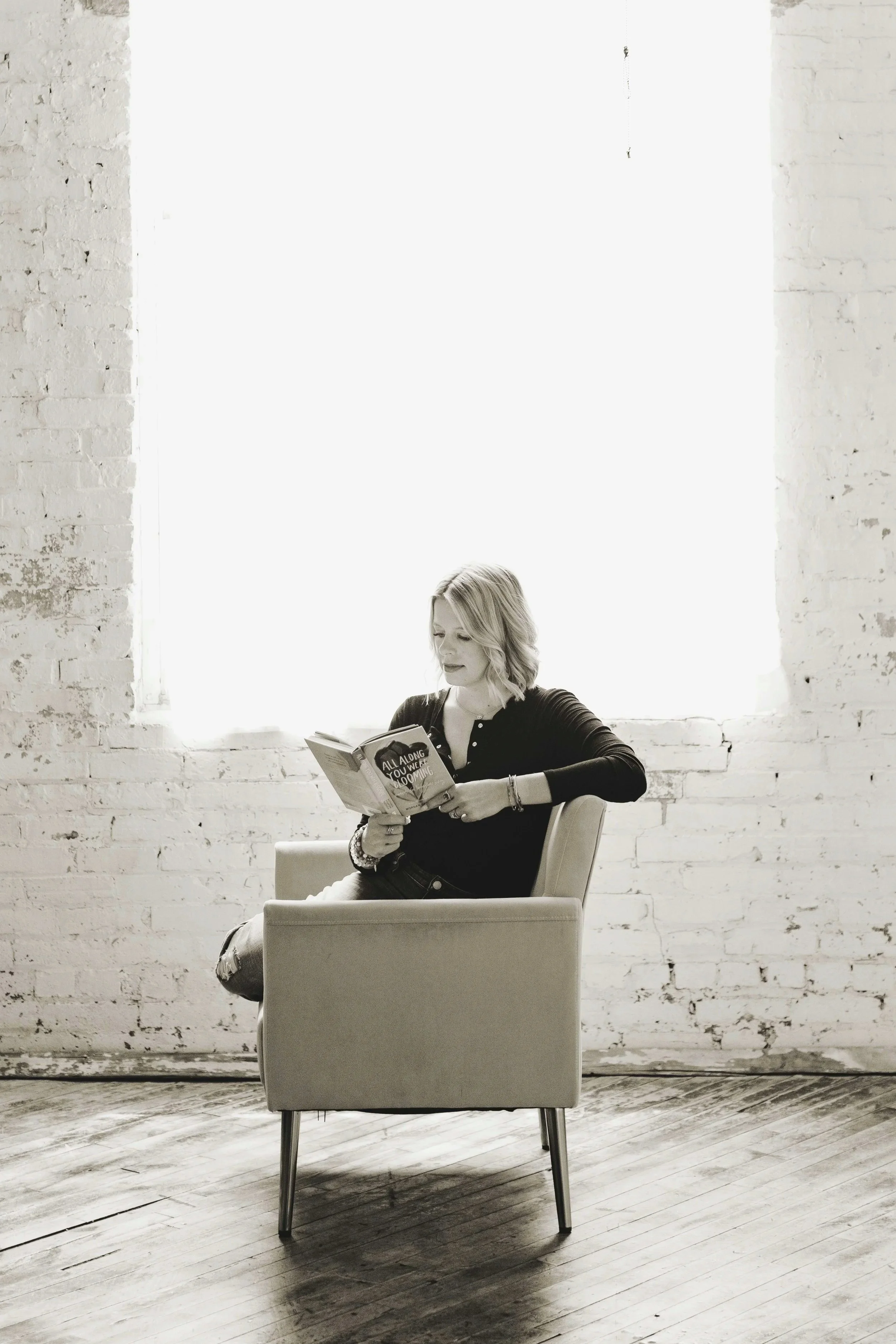 A woman with blonde hair sitting in a beige armchair, reading a book in a room with white brick walls and a large window with bright light coming through.