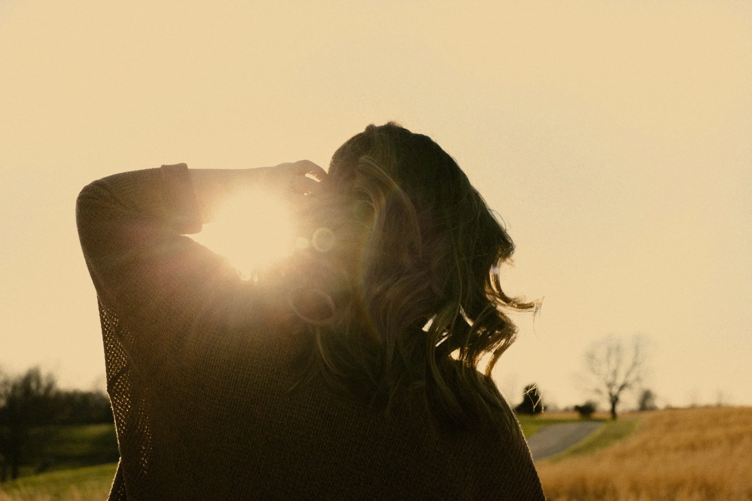A woman with long, wavy hair is outdoors during sunset, with the sun glowing behind her head. She is wearing a mesh top and is holding her hair back with one hand, facing away from the camera. The background features a park with trees and a pathway.