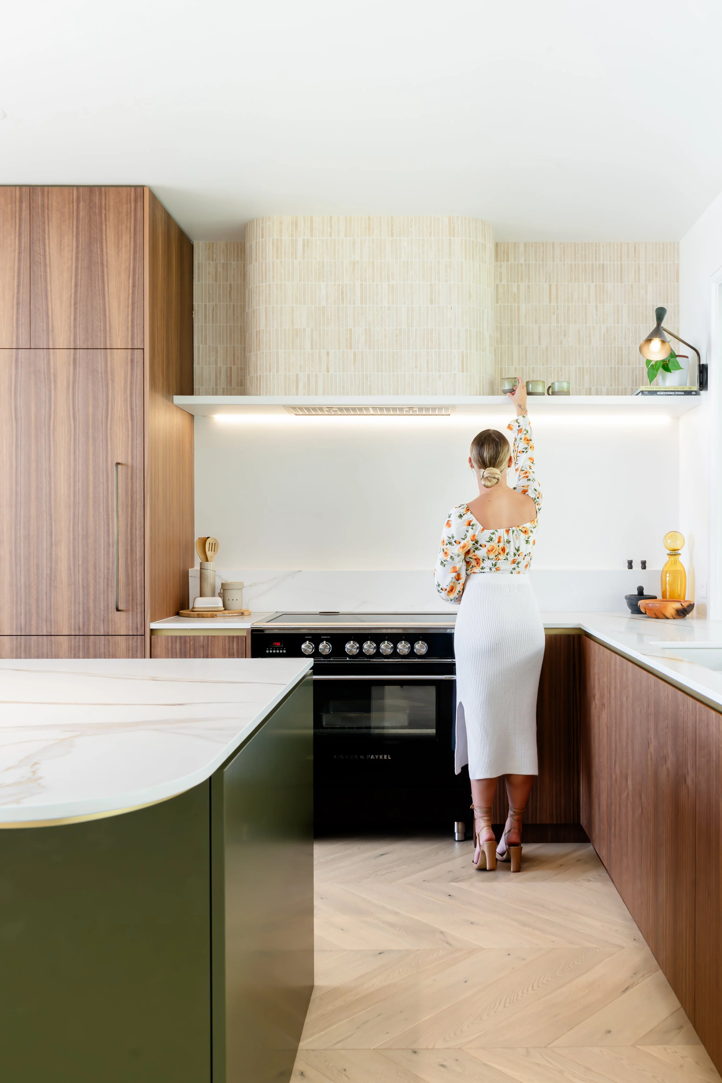 A kitchen completed by Emily which features walnut veneer timber cabinetry, bottle green 2 pac polyurethane, stone benchtops, a curved tiled rangehood, floating shelves and natural lighting.
