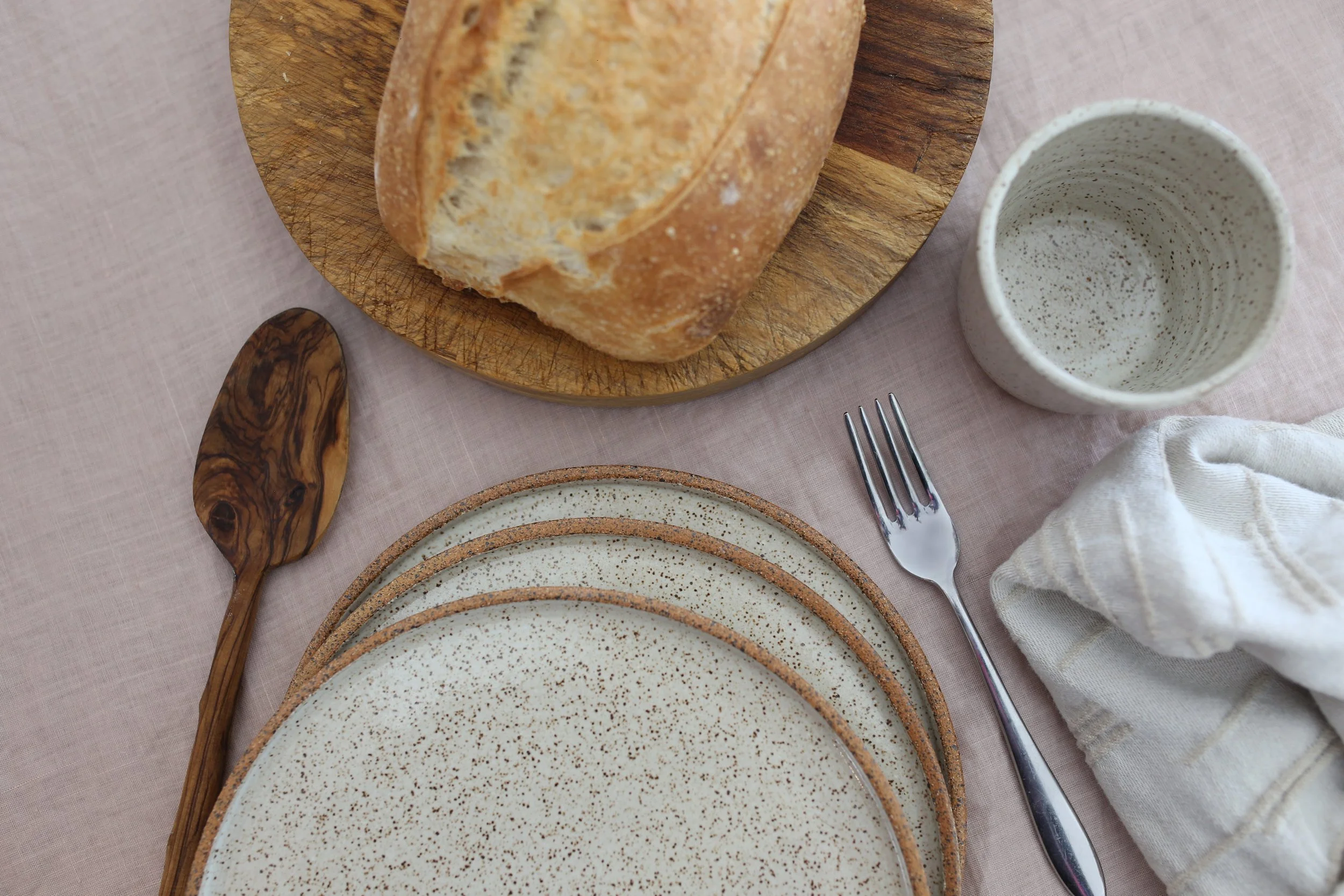 Overhead view of a tableware set up of plates with a loaf of bread on a wooden board, three stacked speckled ceramic plates, a wooden spoon, a speckled mug, a metal fork, and a cloth napkin on a linen tablecloth.