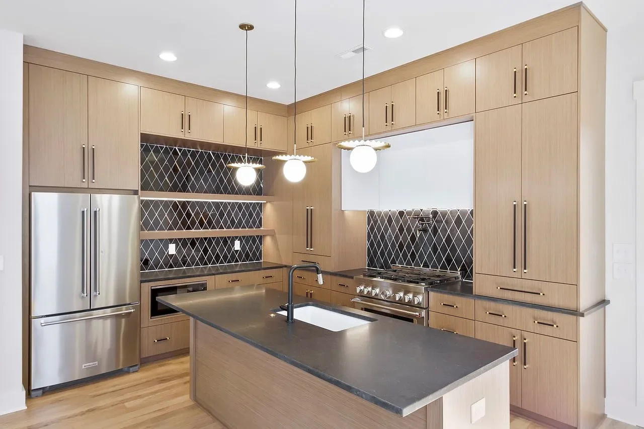 Open-concept kitchen and dining area with gray cabinets, wooden island, dining table, and chairs. Large windows with a view of trees, hardwood floors, and ceiling beams with pendant lights.