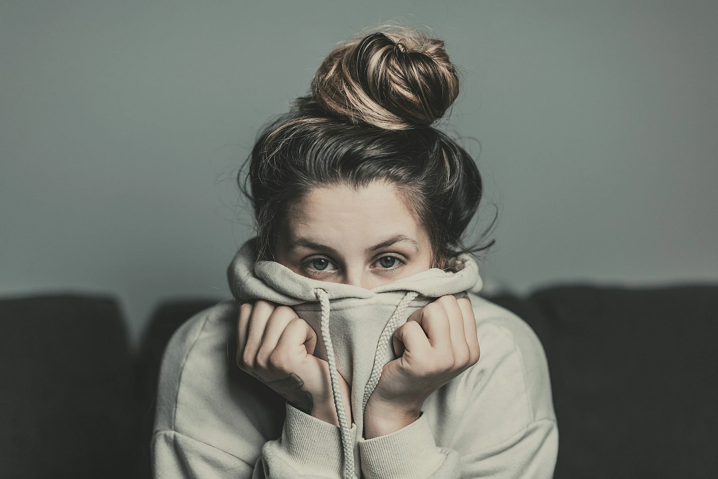 A Caucasian woman with dirty blonde hair up in a messy bun wearing a beige sweatshirt covering all of her face except her eyes appearing stressed symbolizing a need for anxiety therapy in Connecticut