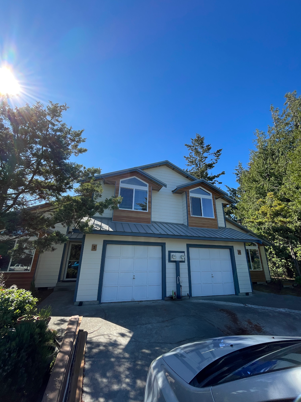 Two-story house with a double garage, light-colored siding, and windows, surrounded by trees under a clear blue sky, sunlight shining from the upper left.