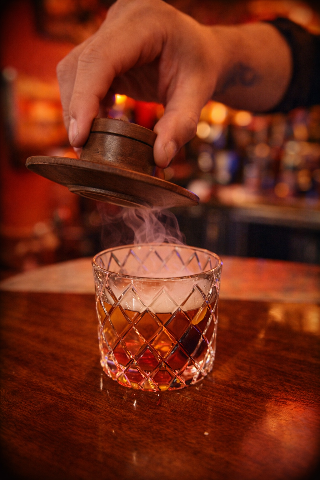 A hand holding a wooden disc-shaped object above a glass filled with a dark amber liquid, with vapor rising from the drink, on a wooden bar counter with warm ambient lighting.