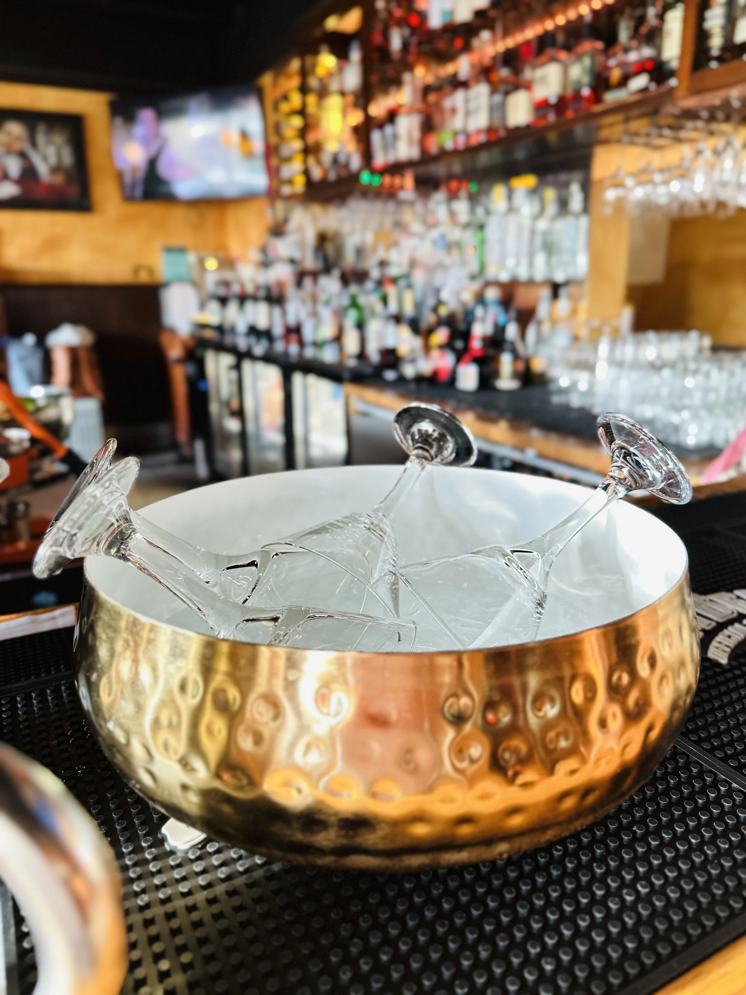 A shiny metal bowl filled with ice and upside-down wine glasses on a bar counter. The background shows a blurry bar with bottles, glasses, and a TV.