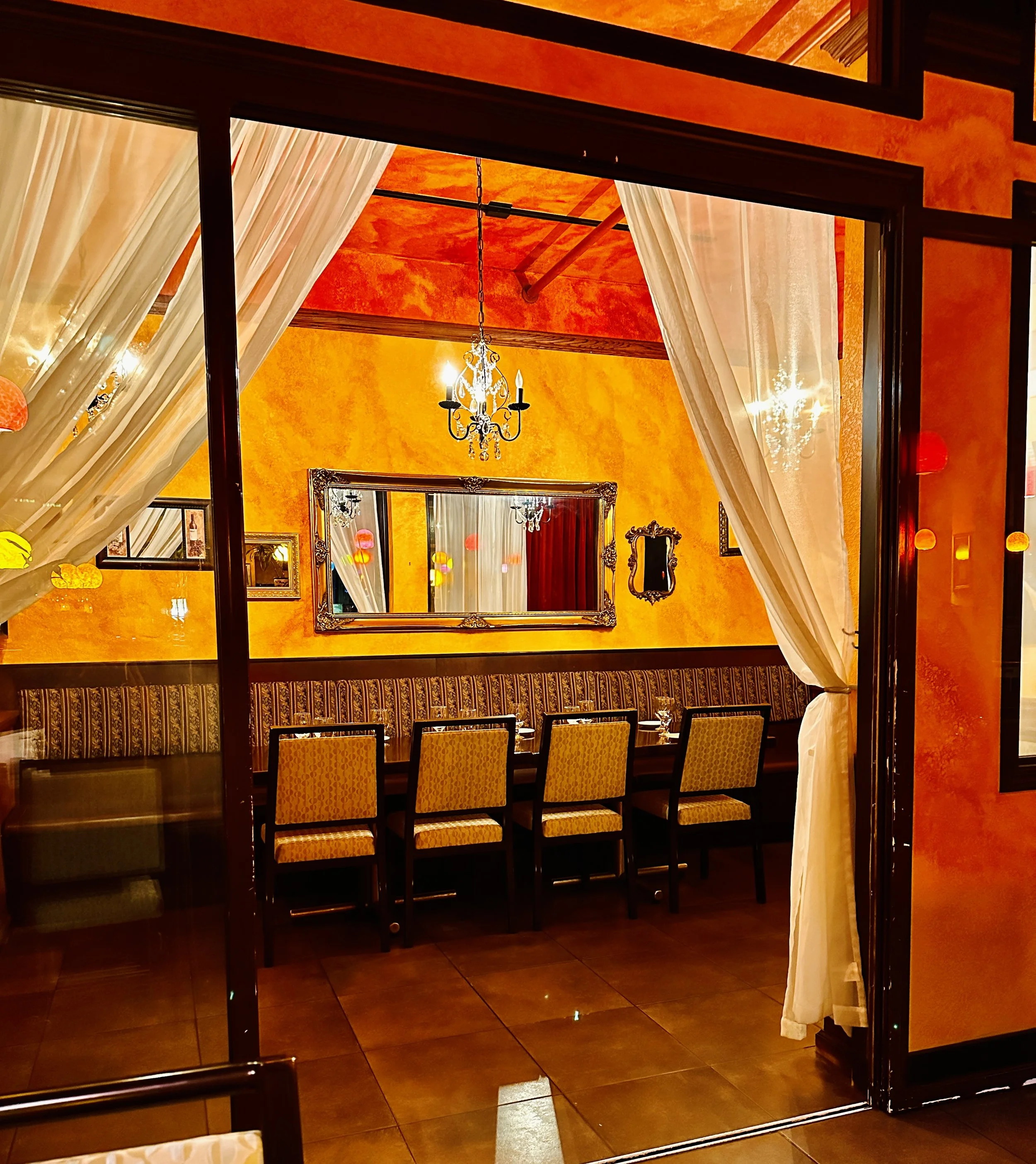 Elegant restaurant with orange textured walls, a chandelier, a mirror, and six patterned chairs along a long table, viewed through a doorway with white curtains.