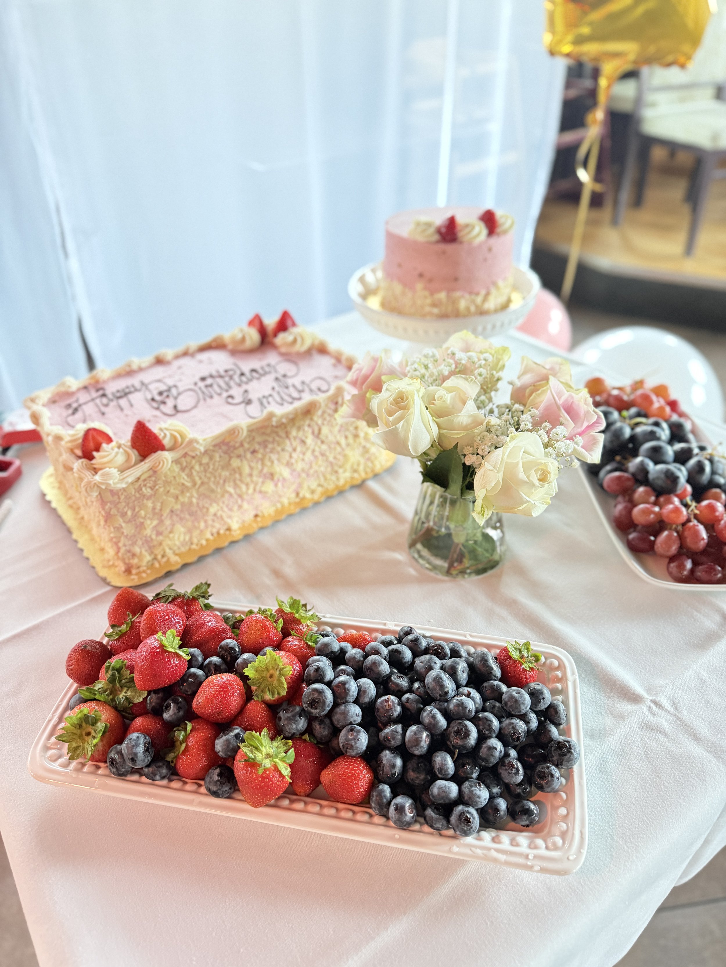A table decorated with strawberries and blueberries, a bouquet of pink and white roses, and two pink birthday cakes with cream and strawberries, celebrating a birthday.