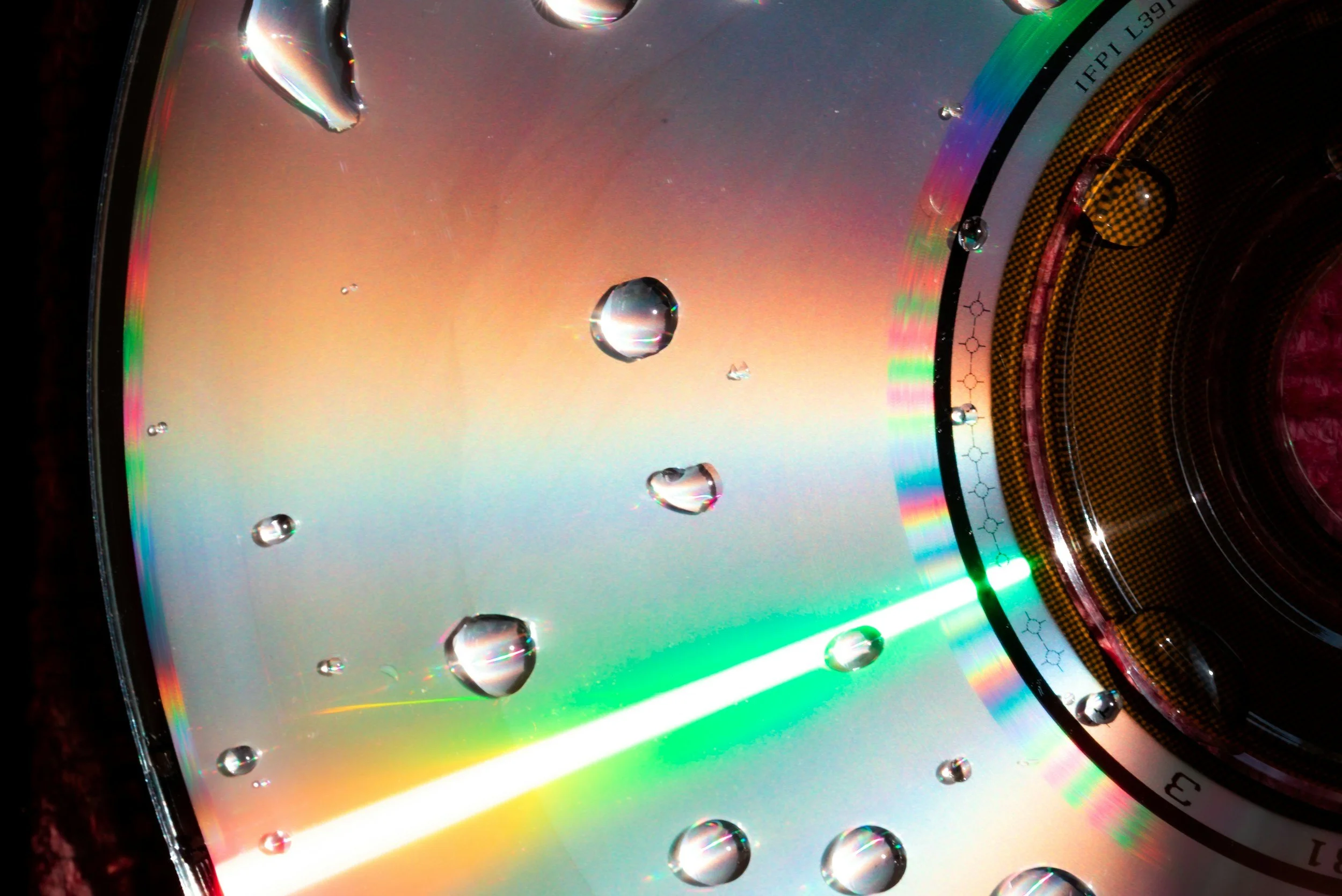 Close-up of an inner surface of a CD with water droplets on it, showing rainbow colors and a reflective surface.